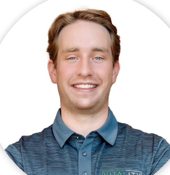 A young man with light brown hair and blue eyes smiling, wearing a blue collared shirt with a name tag that reads "VITALITY."