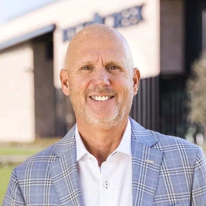 A smiling middle-aged man with a bald head, light beard, and light skin, wearing a gray plaid blazer and a white shirt, standing outdoors near a building.