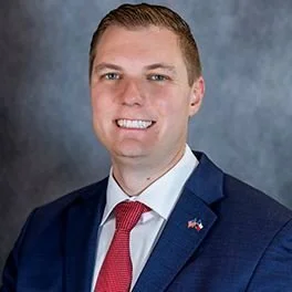 Portrait of a man in a dark blue suit, white shirt, and red tie, smiling, against a gray background.
