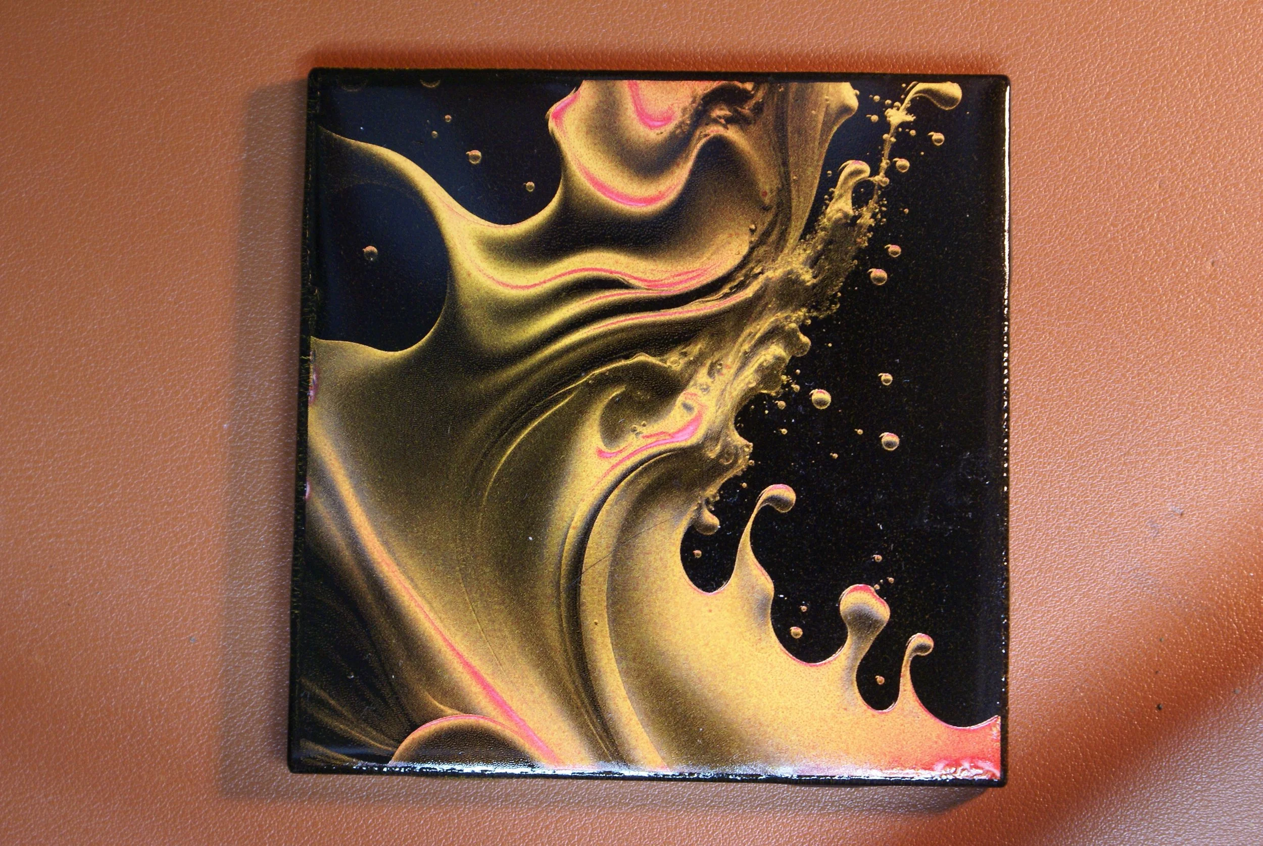 An abstract painting with swirling black, gold, and pink colors on a square canvas placed on a brown surface.