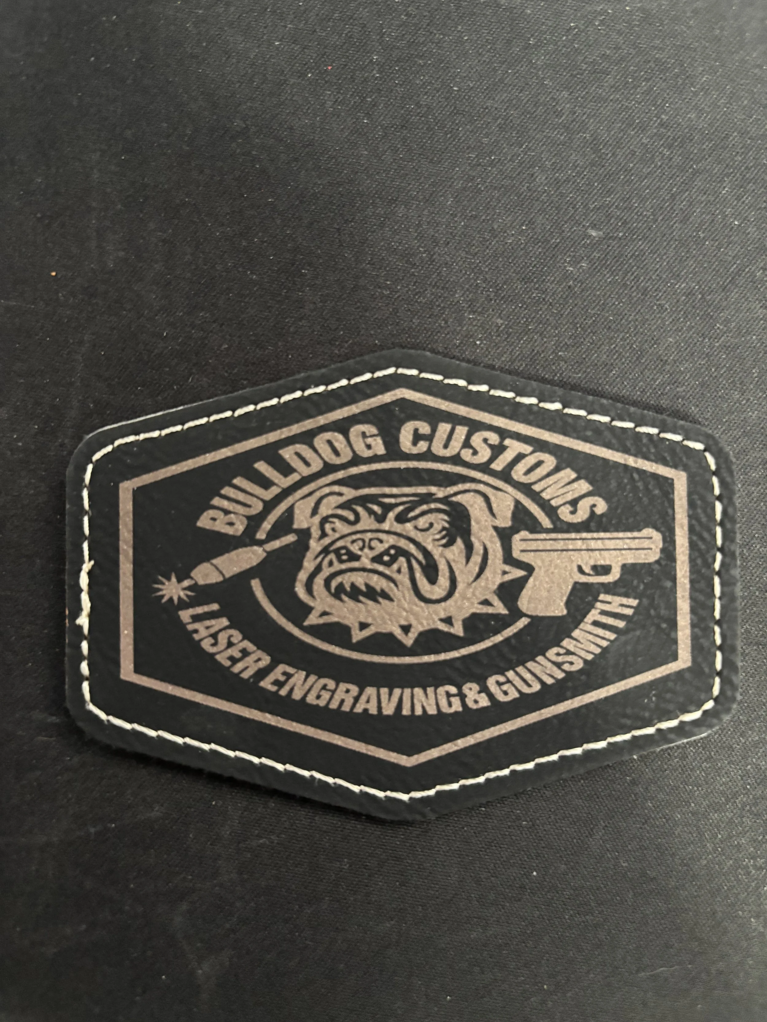 A black patch with beige stitching and text that reads "Bulldog Customs Laser Engraving & Gunsmithing". It features an image of an angry bulldog with a collar, a laser, and a gun.