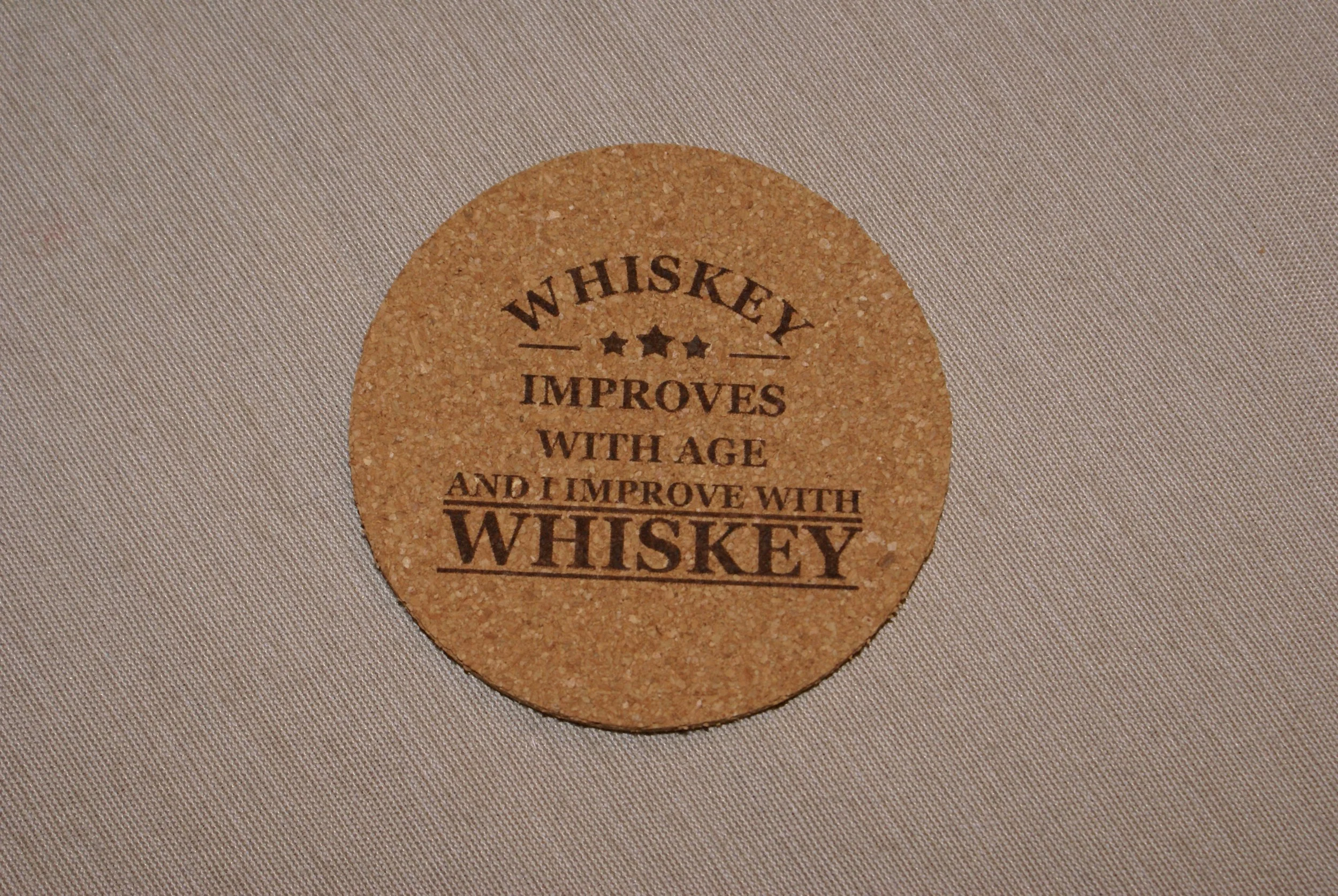 A round cork coaster with the text 'Whiskey improves with age and I improve with whiskey,' printed in black on it, placed on a beige fabric surface.