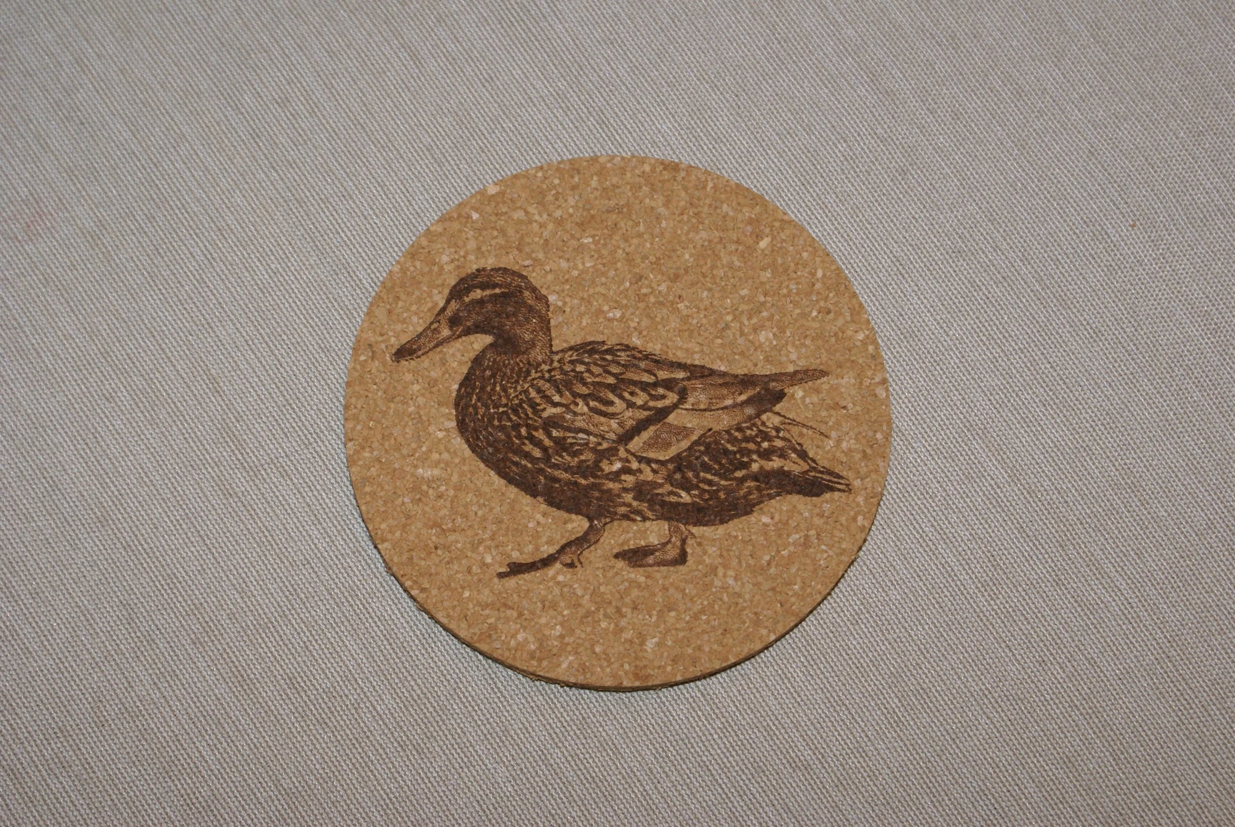 A round cork coaster with a detailed illustration of a duck in dark brown.