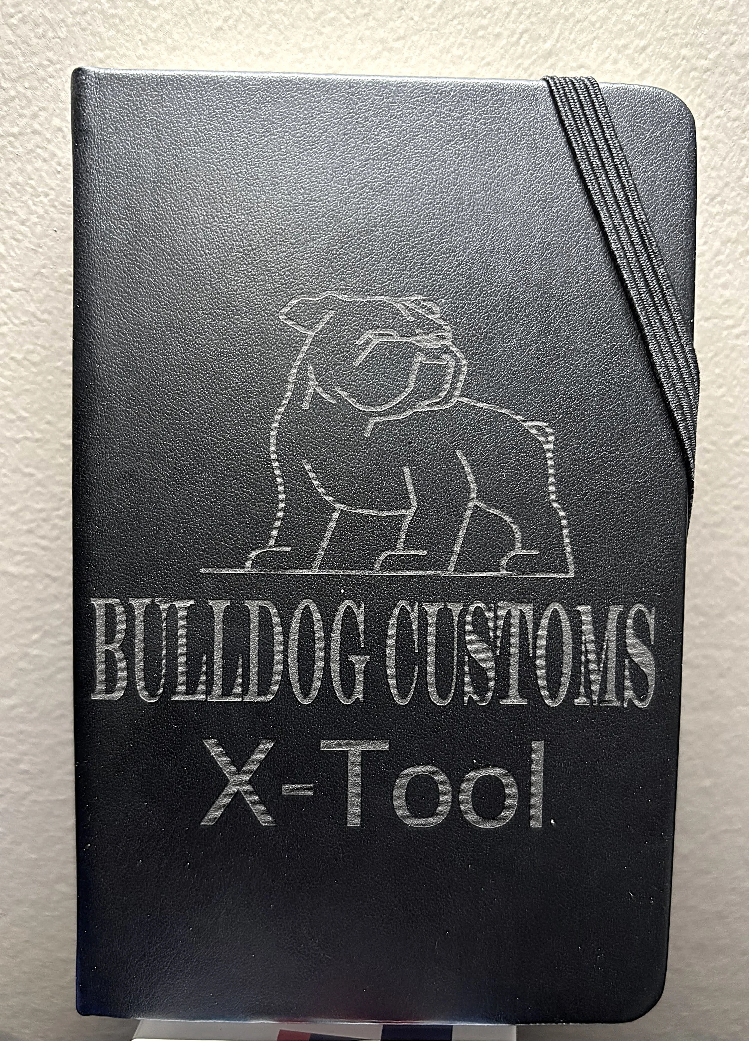 A black notebook with a dog illustration and the words 'BULLDOG CUSTOMS X-Tool' embossed on the cover.