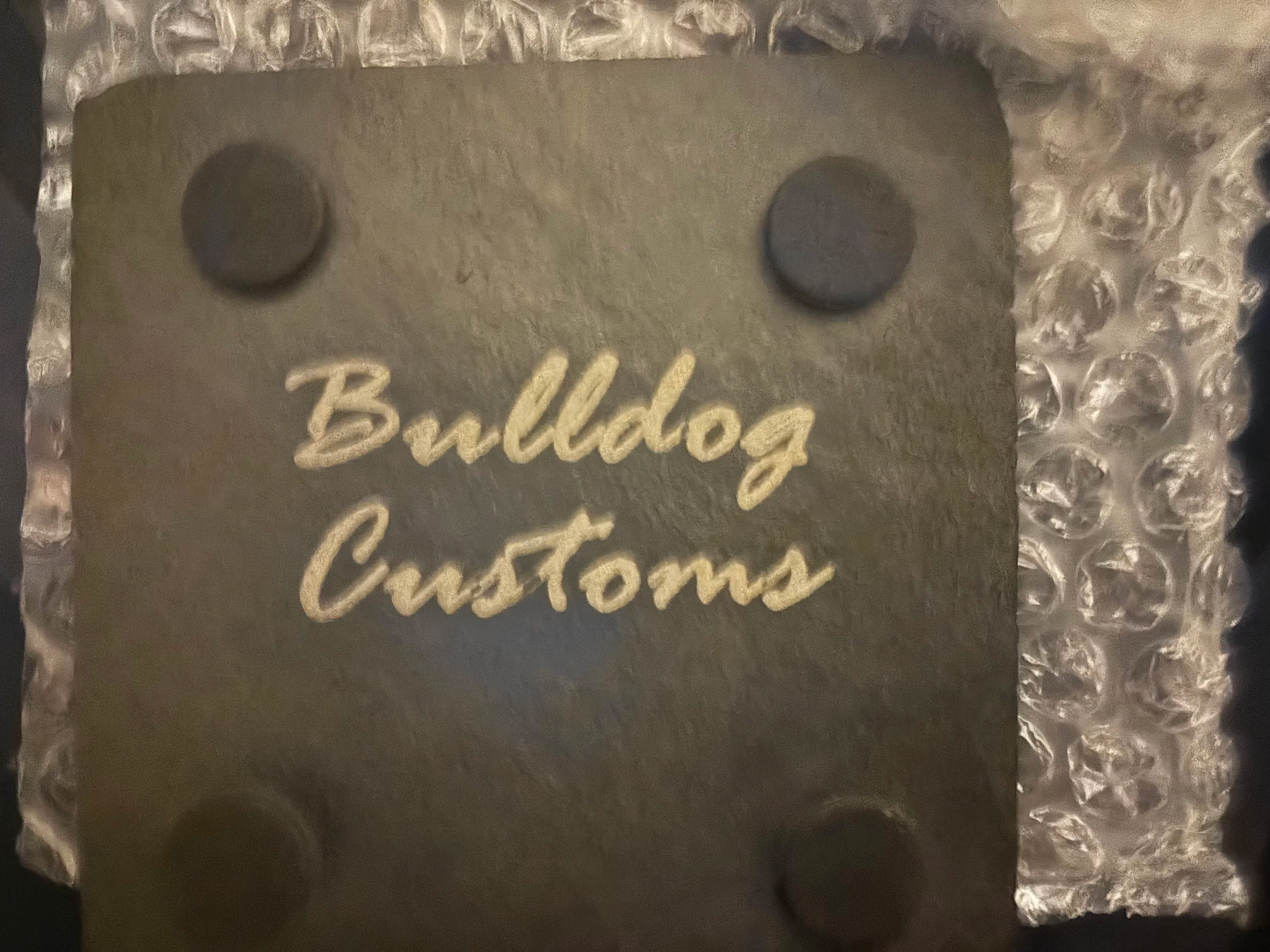 Close-up of a dark-colored rectangular object with the words 'Bulldog Customs' written in light-colored script, secured to a textured metallic surface with four round fasteners, one at each corner.
