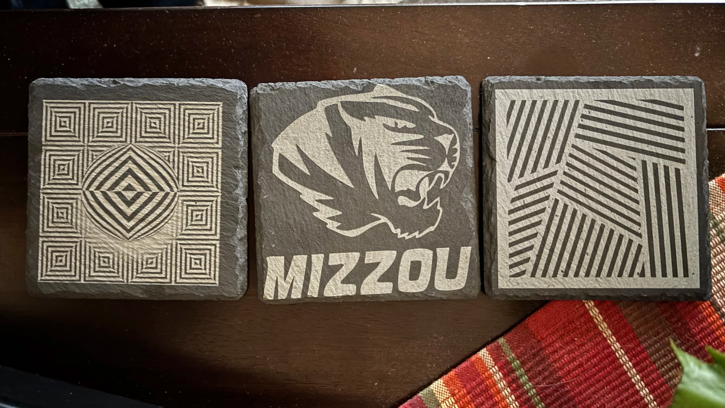 Three decorative stone tiles placed on a wooden surface. The first tile has a geometric pattern with concentric diamonds and squares. The second tile features a stylized roaring lion's head with the word 'MIZZOU' beneath it. The third tile displays a