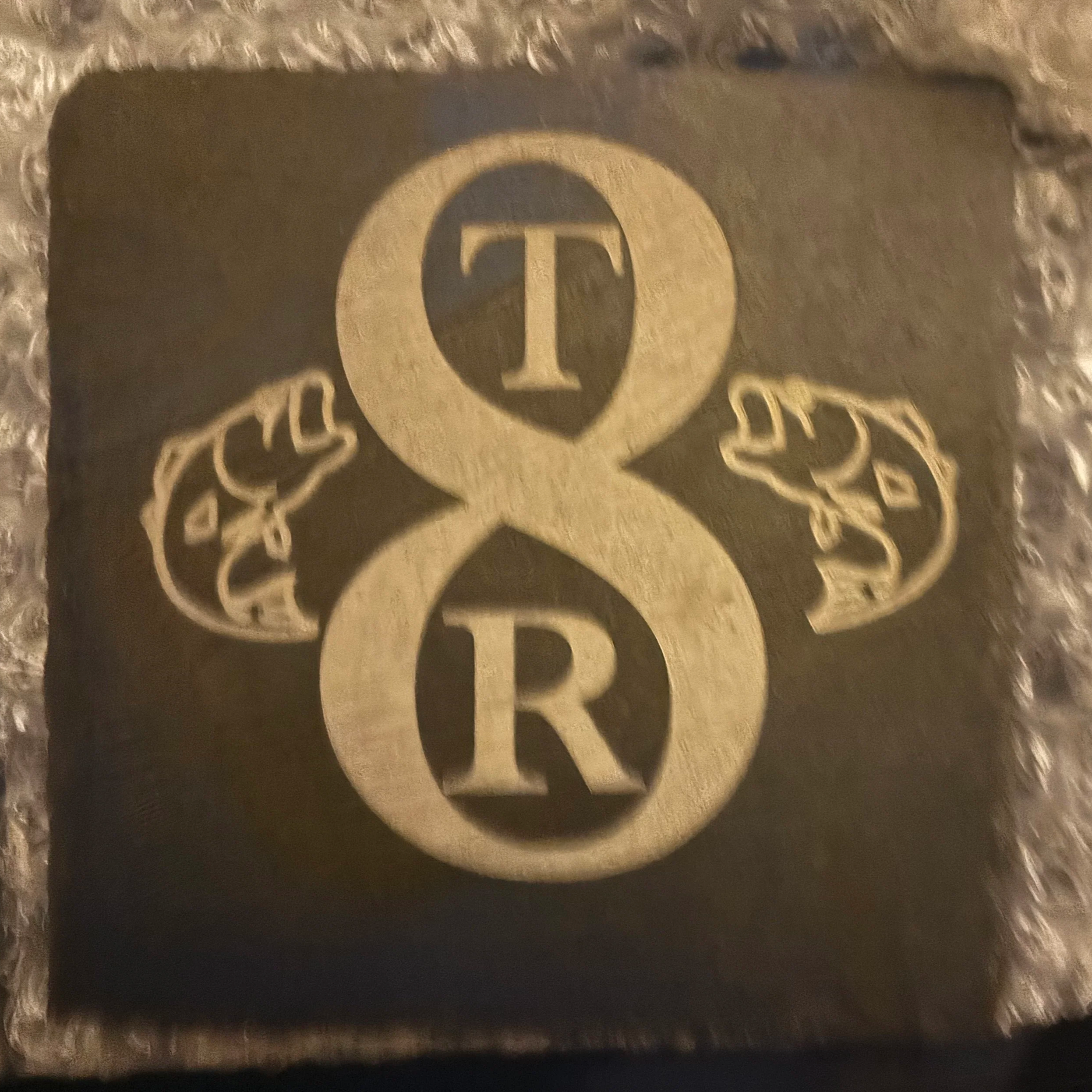 A metal pendant with a design featuring two vertically stacked circles with the letters 'T' and 'R' inside them, connected by a stylized ampersand. There are two hands reaching towards each other on either side of the design.