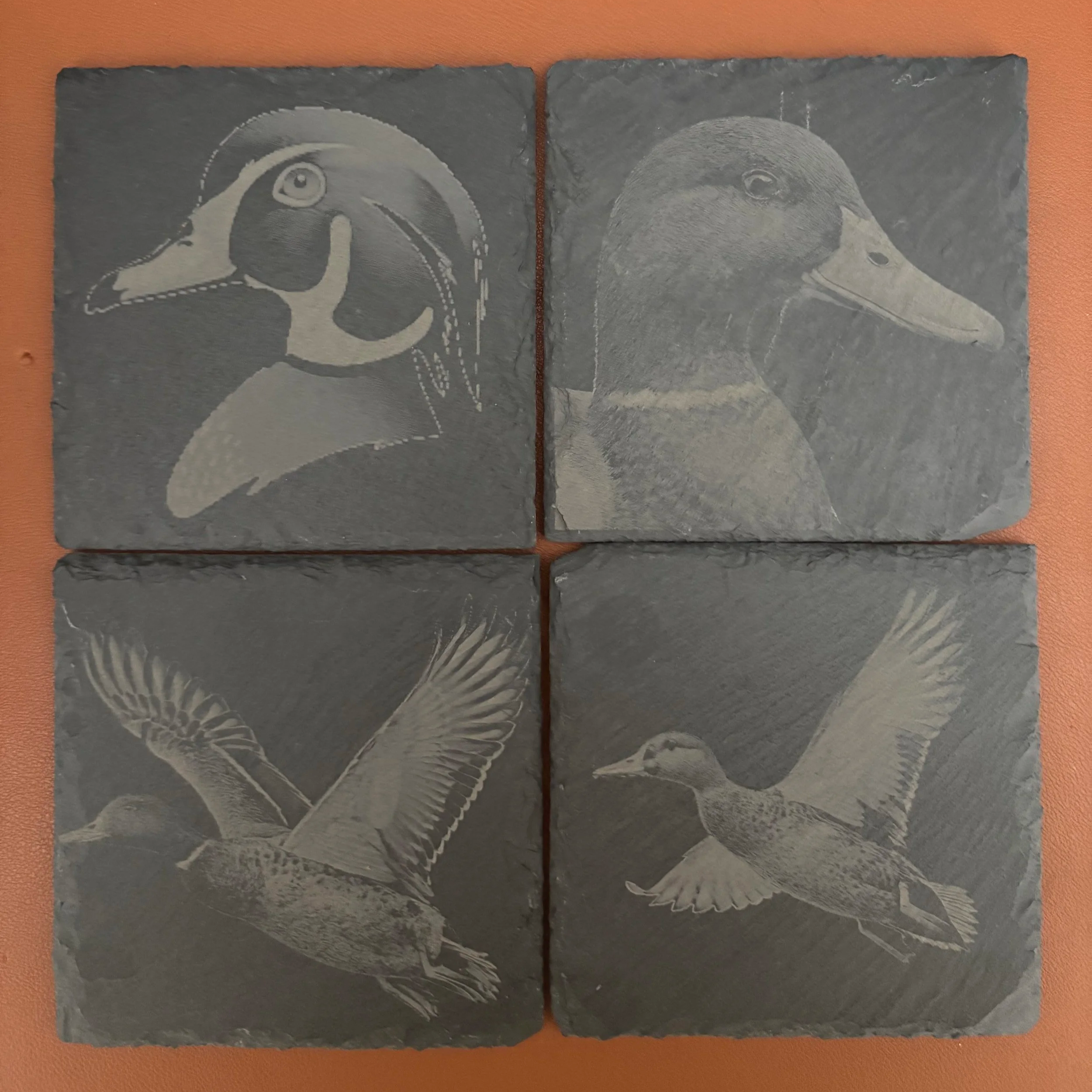 Four slate tiles with etched images of different ducks and waterfowl: two close-up profile views of duck heads at the top, and two flying ducks with wings spread at the bottom.