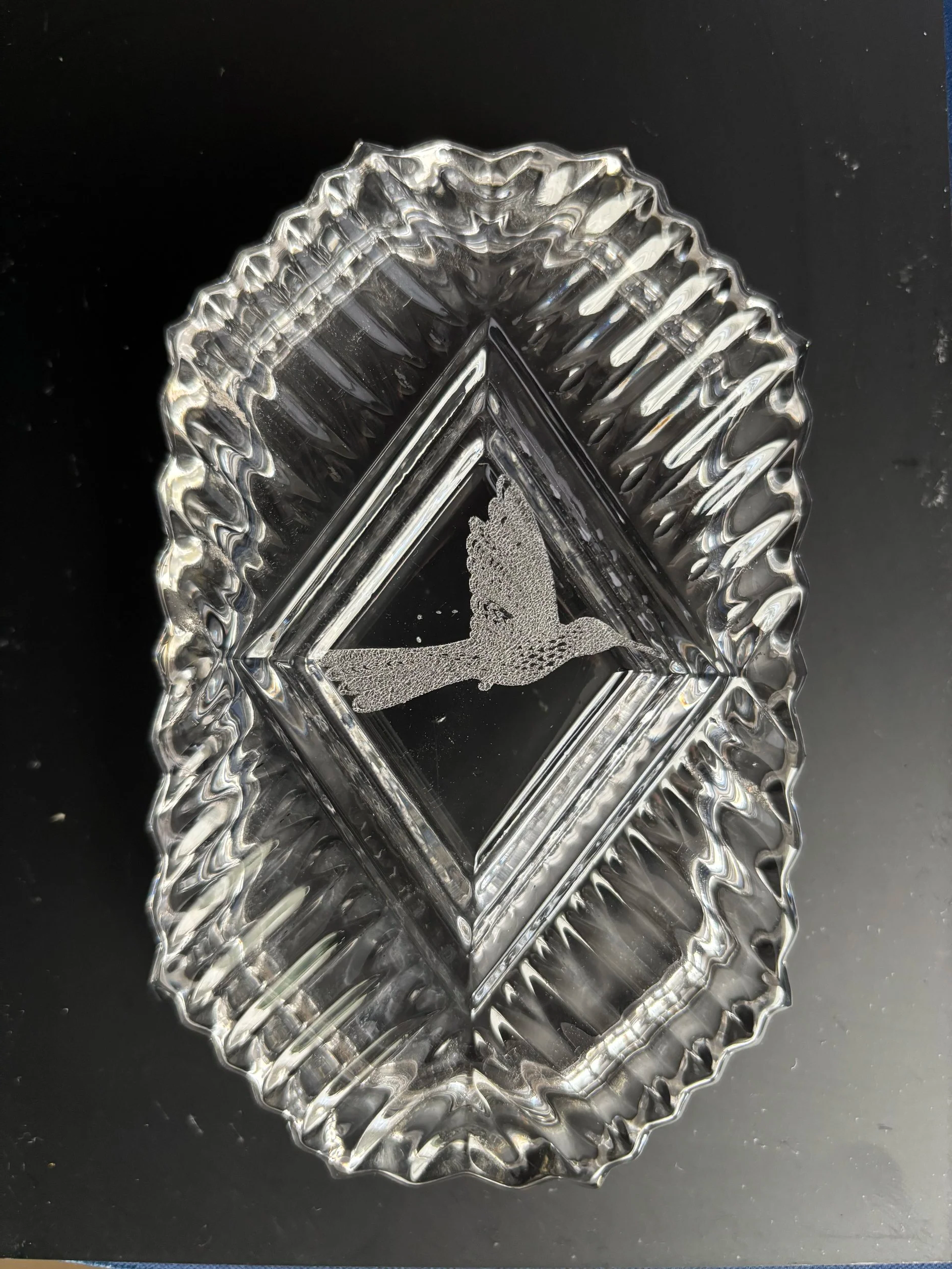 A clear glass dish with a scalloped edge and a diamond-shaped interior, featuring a 3D hummingbird figurine inside, against a black background.
