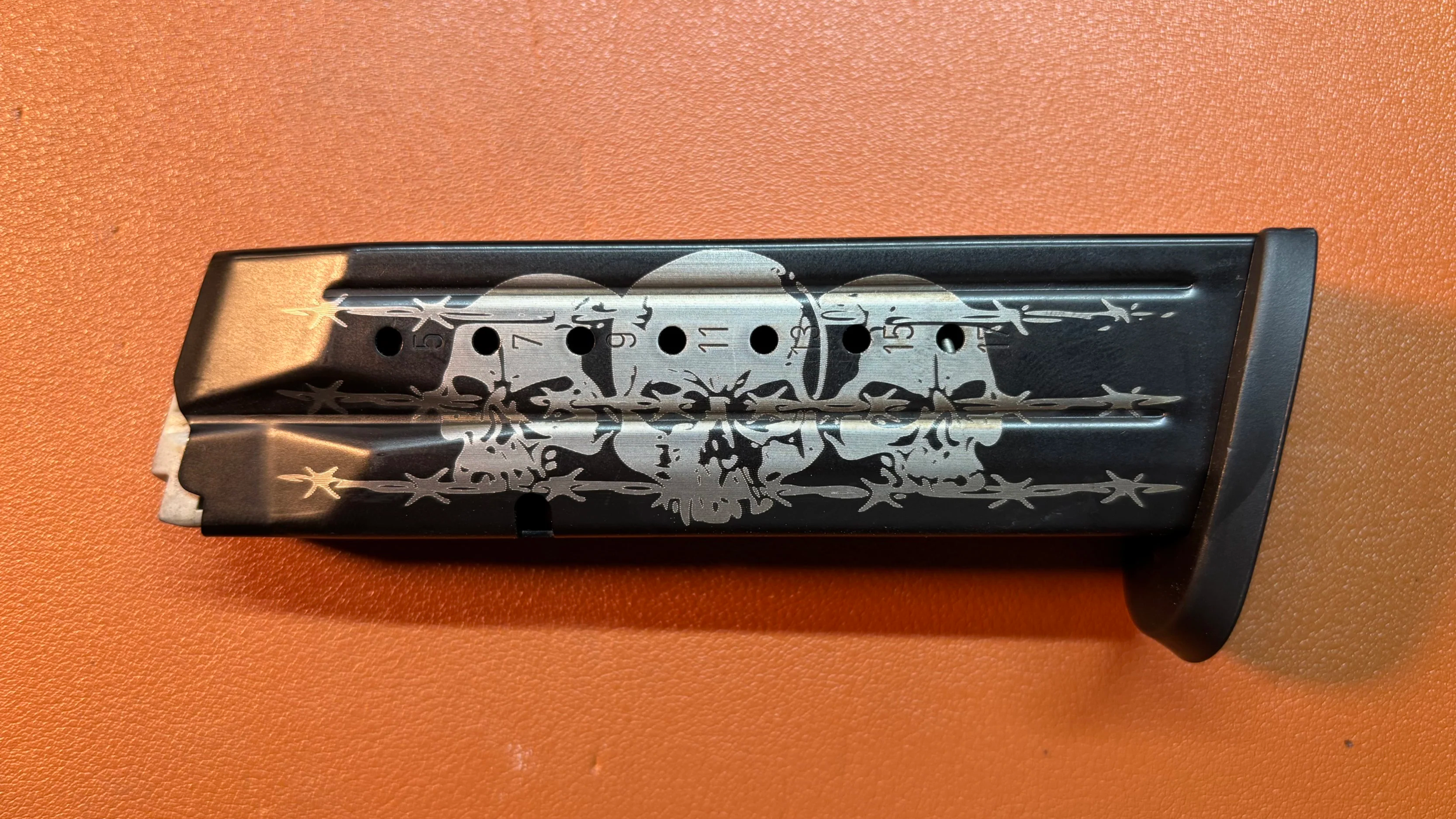 A black skateboard deck with a white skull and floral design, and numbered holes for mounting trucks, on an orange textured surface.