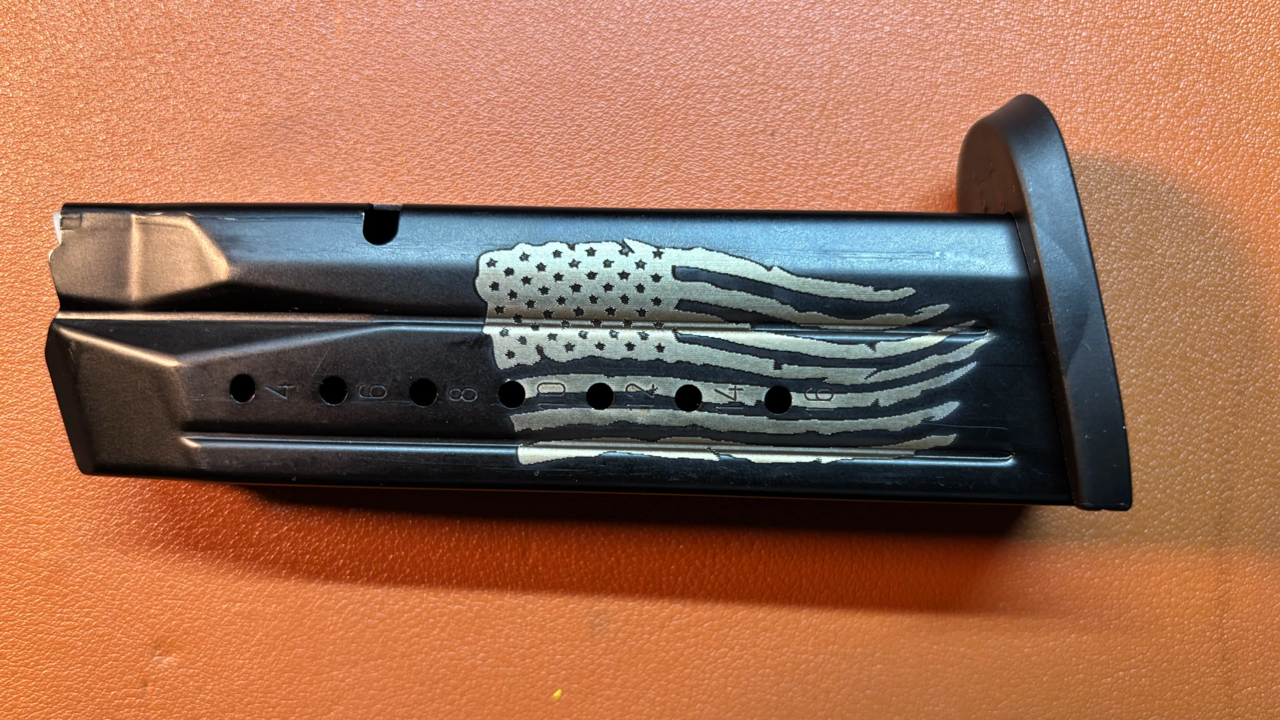 A detached black magazine with a silver American flag design on the side, placed on an orange surface.
