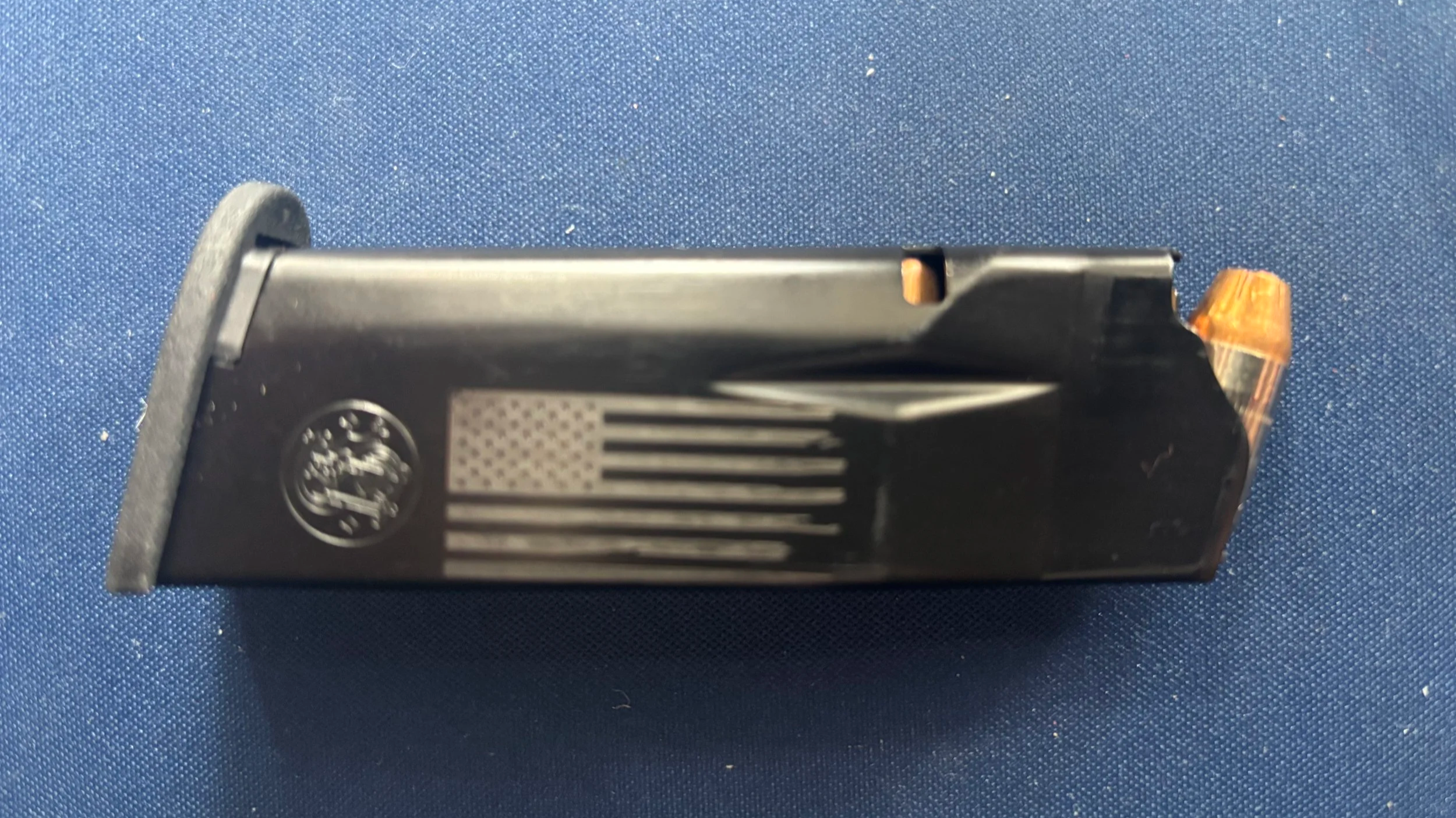 A black semi-automatic handgun with a magazine inserted, featuring an American flag and circular emblem on the side, placed on a textured blue surface.