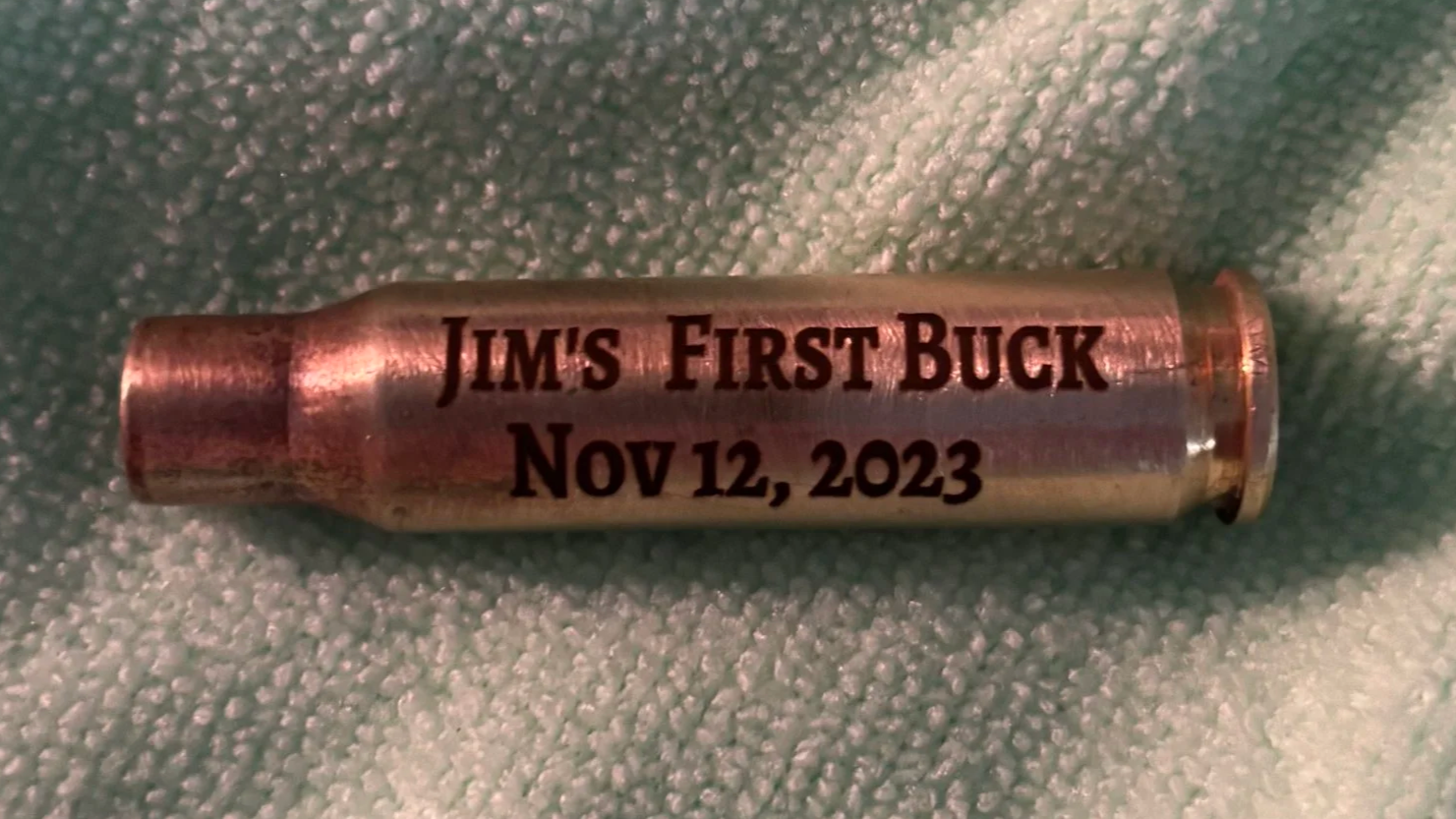 A metallic shell casing with black text reading 'Jim's First Buck Nov 12, 2023' on a soft textured surface.