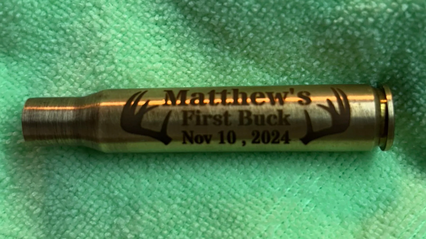 A metallic shotgun shell with black text and a graphic of deer antlers, personalized with the name 'Matthew's', the words 'First Buck', and the date 'Nov 10, 2024', resting on a soft, textured green fabric background.