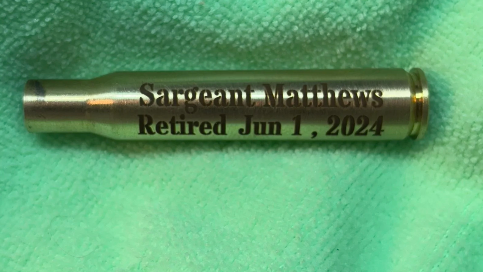 Metal tube with engraved text reading "Sargeant Matthews Retired Jun 1, 2024" on a green cloth background.