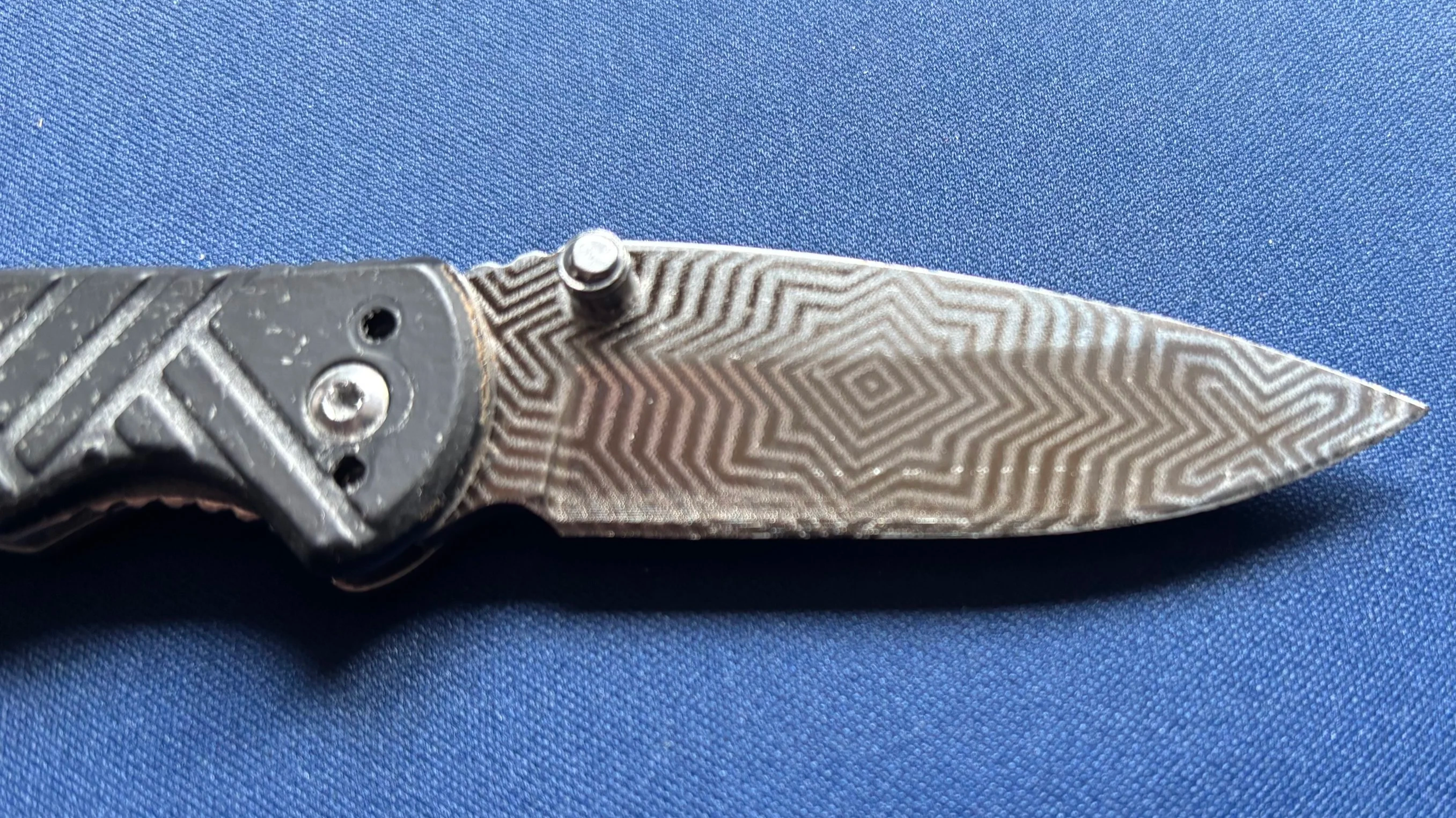 Close-up of a folding knife with a patterned metal blade and a black handle, resting on a blue fabric background.
