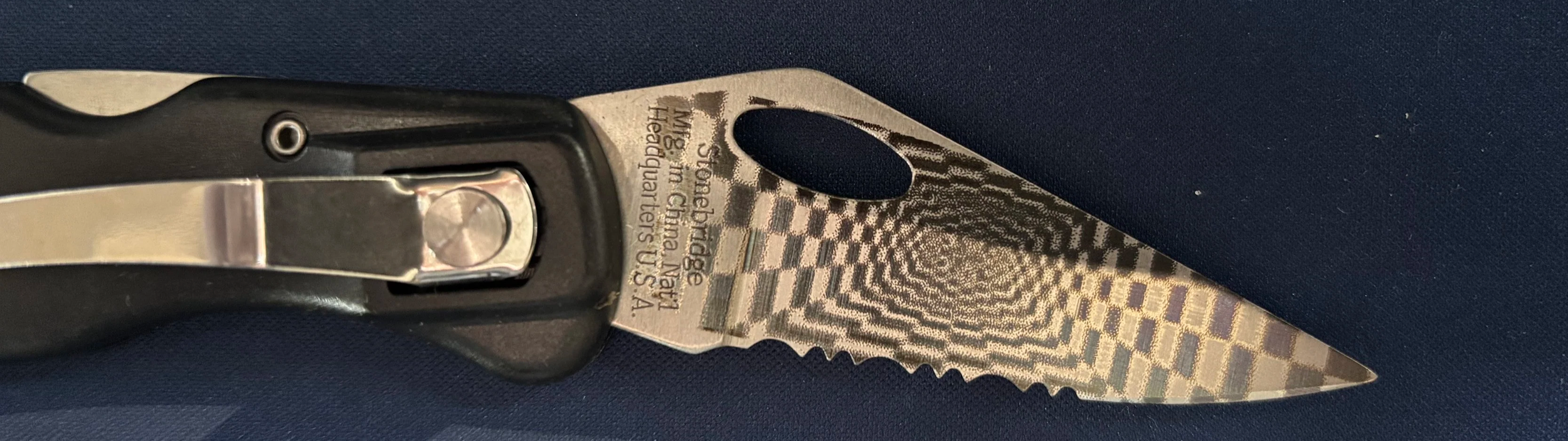 Close-up of a folding pocket knife with a patterned blade and black handle, resting on a dark blue surface.