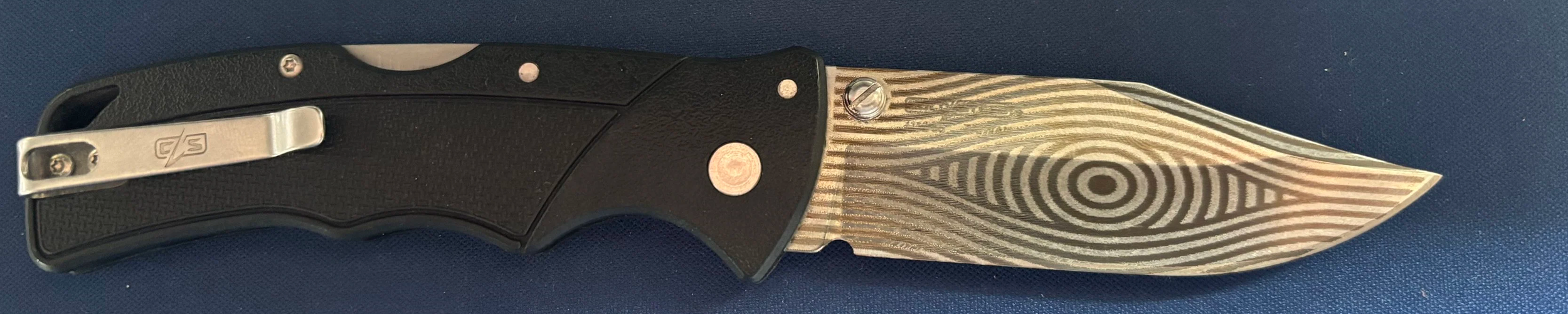 A folding pocket knife with a black handle and a patterned metallic blade.