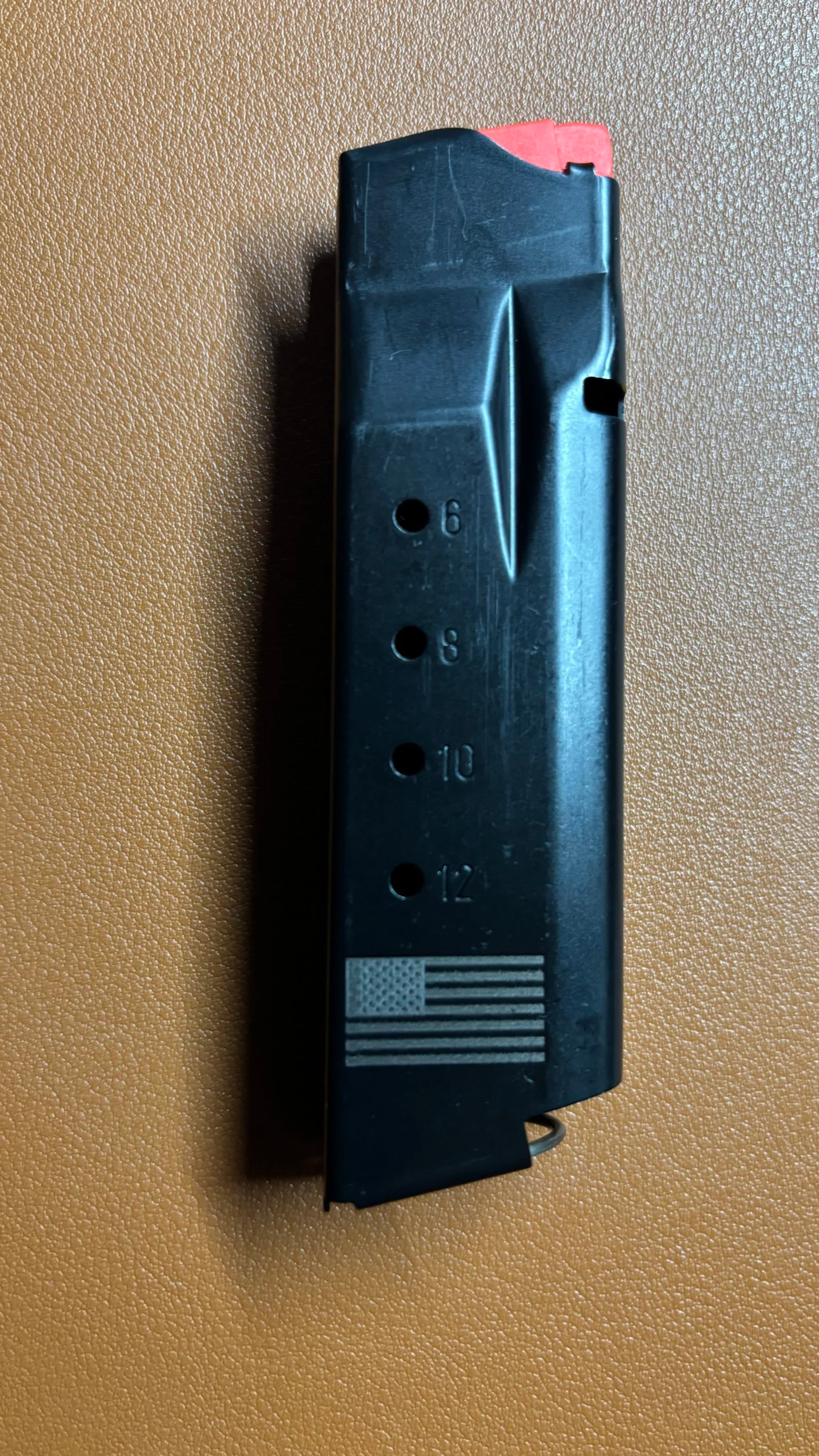 A black firearm magazine with a red top, placed on a textured brown surface, marked with caliber sizes 9mm, 6mm, 10mm, and 12mm, and an American flag graphic.