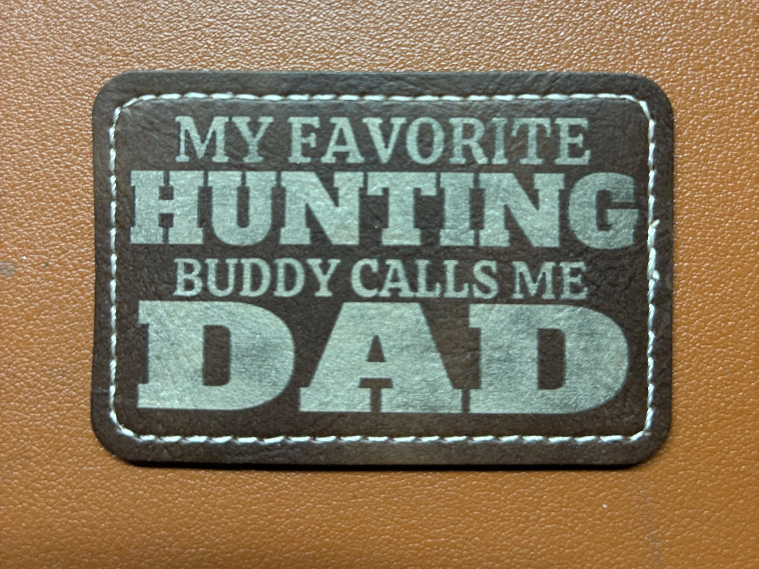 A patch with the text 'My Favorite Hunting Buddy Calls Me Dad' sewn on a brown surface.