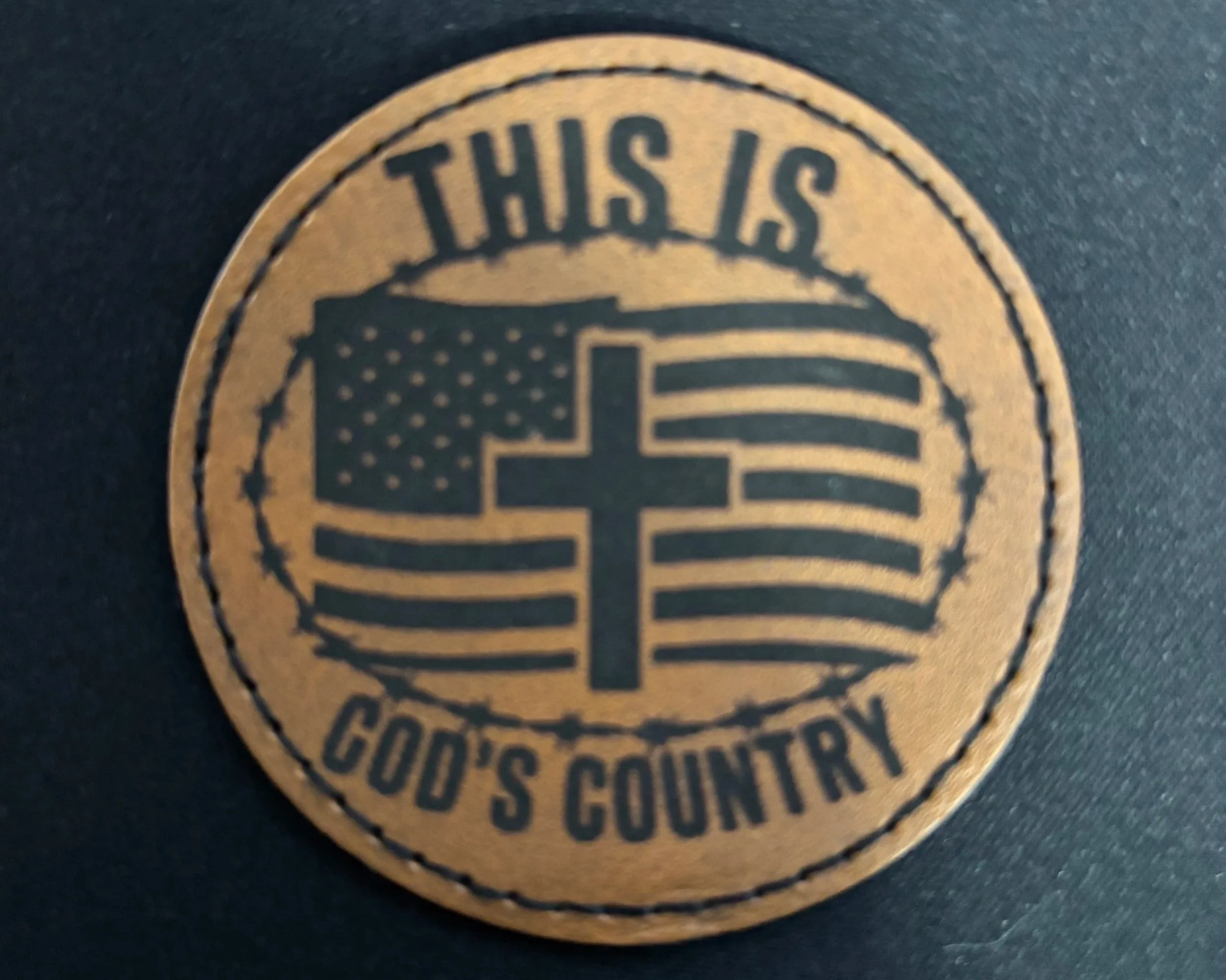 A circular leather patch with the text "This is God's Country" surrounding a graphic of the American flag with a cross at the center.