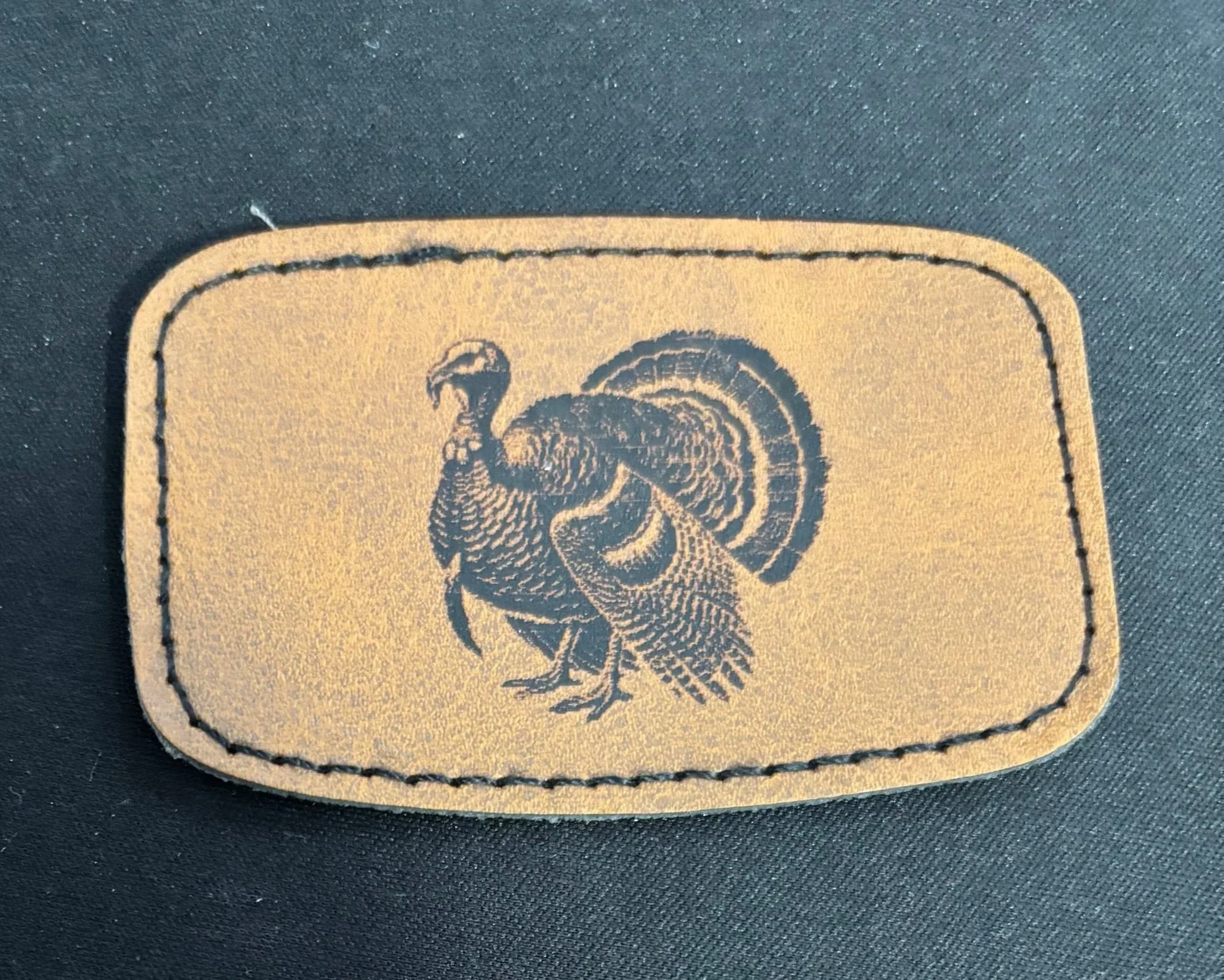 A beige rectangular patch with a black embroidered turkey design and a stitched border, placed on a dark fabric background.