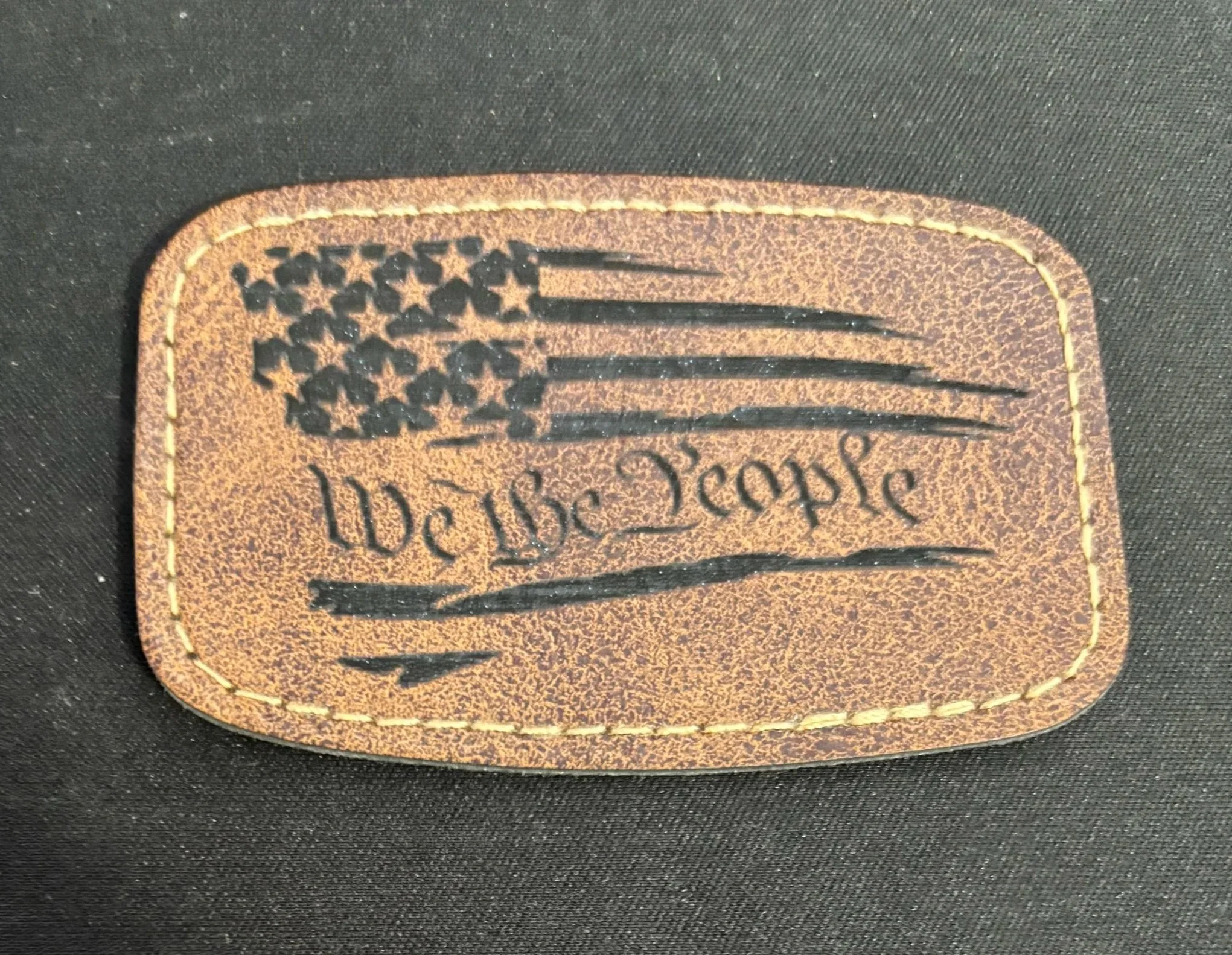 A leather patch featuring an American flag with stars and stripes, the words 'We The People', and a stitched border, placed on a dark fabric background.