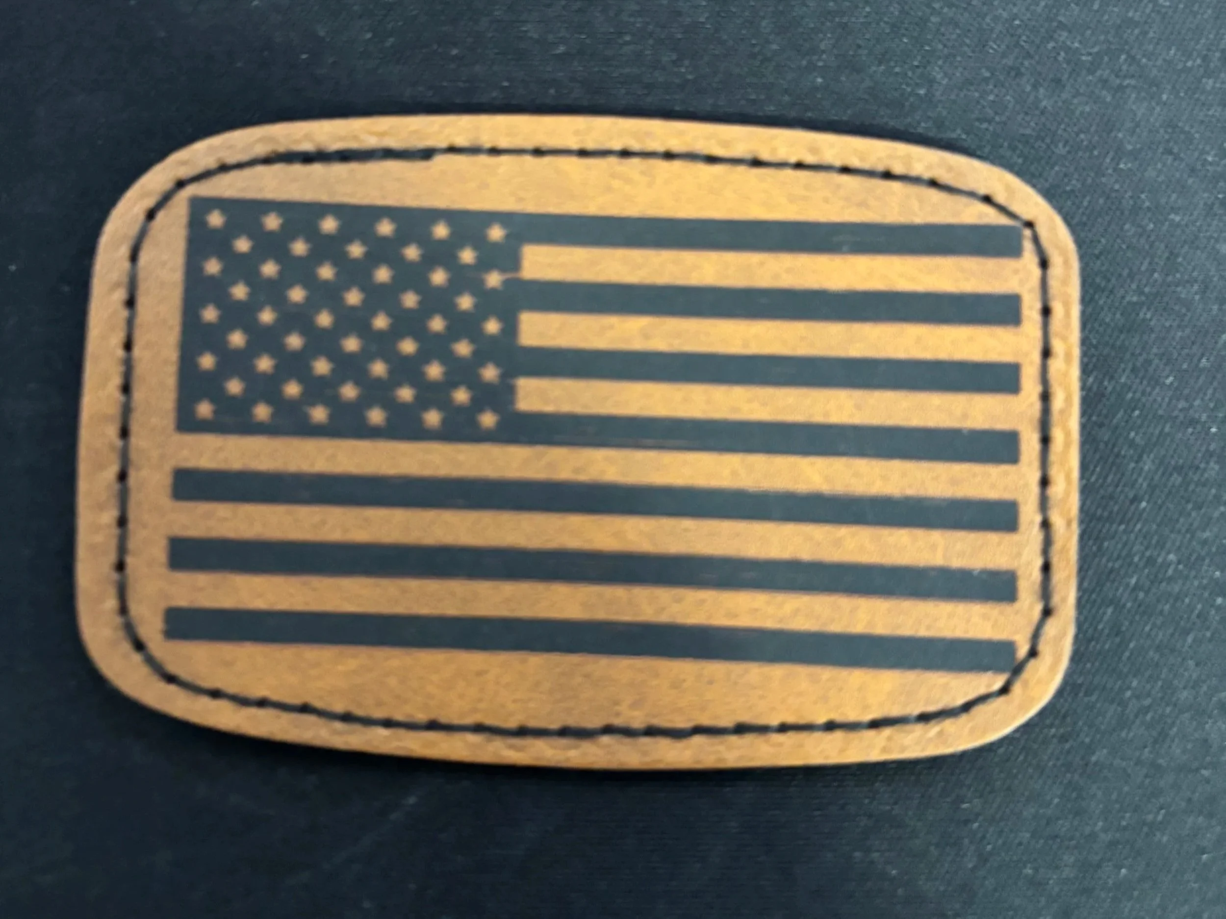 A patch with a stylized American flag design, featuring stars and stripes in a monochromatic color scheme, sewn onto a dark fabric background.
