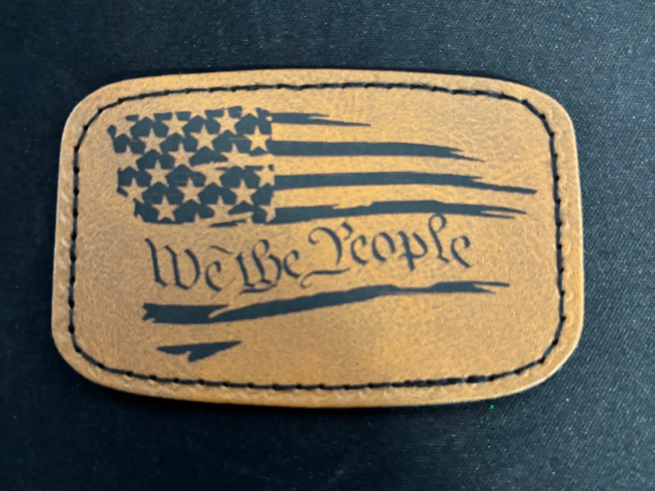 Tan leather patch with black printed American flag and the words "We the People" underneath.