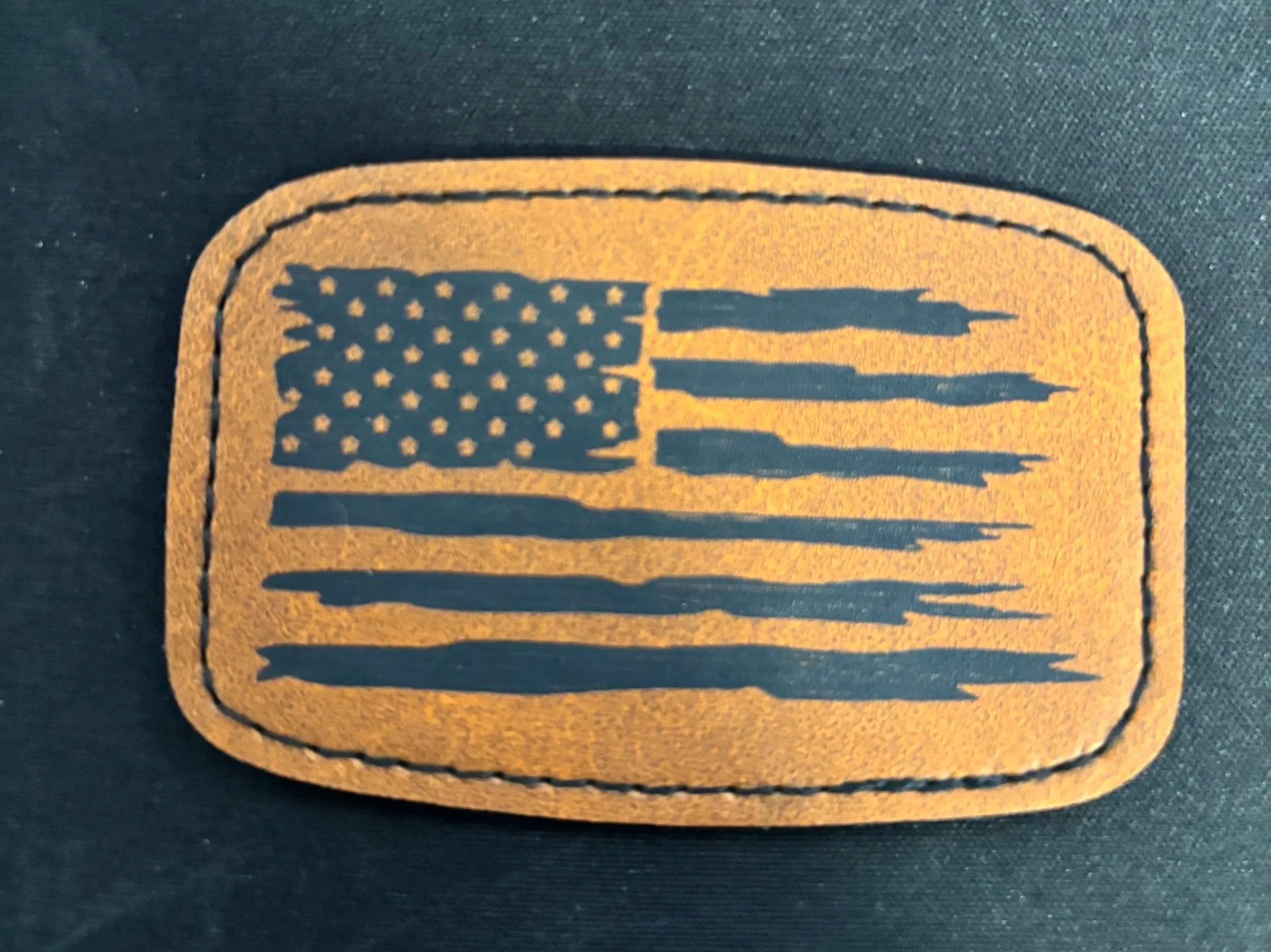 A leather patch featuring a stylized American flag with black and gold colors, stitched along the border.