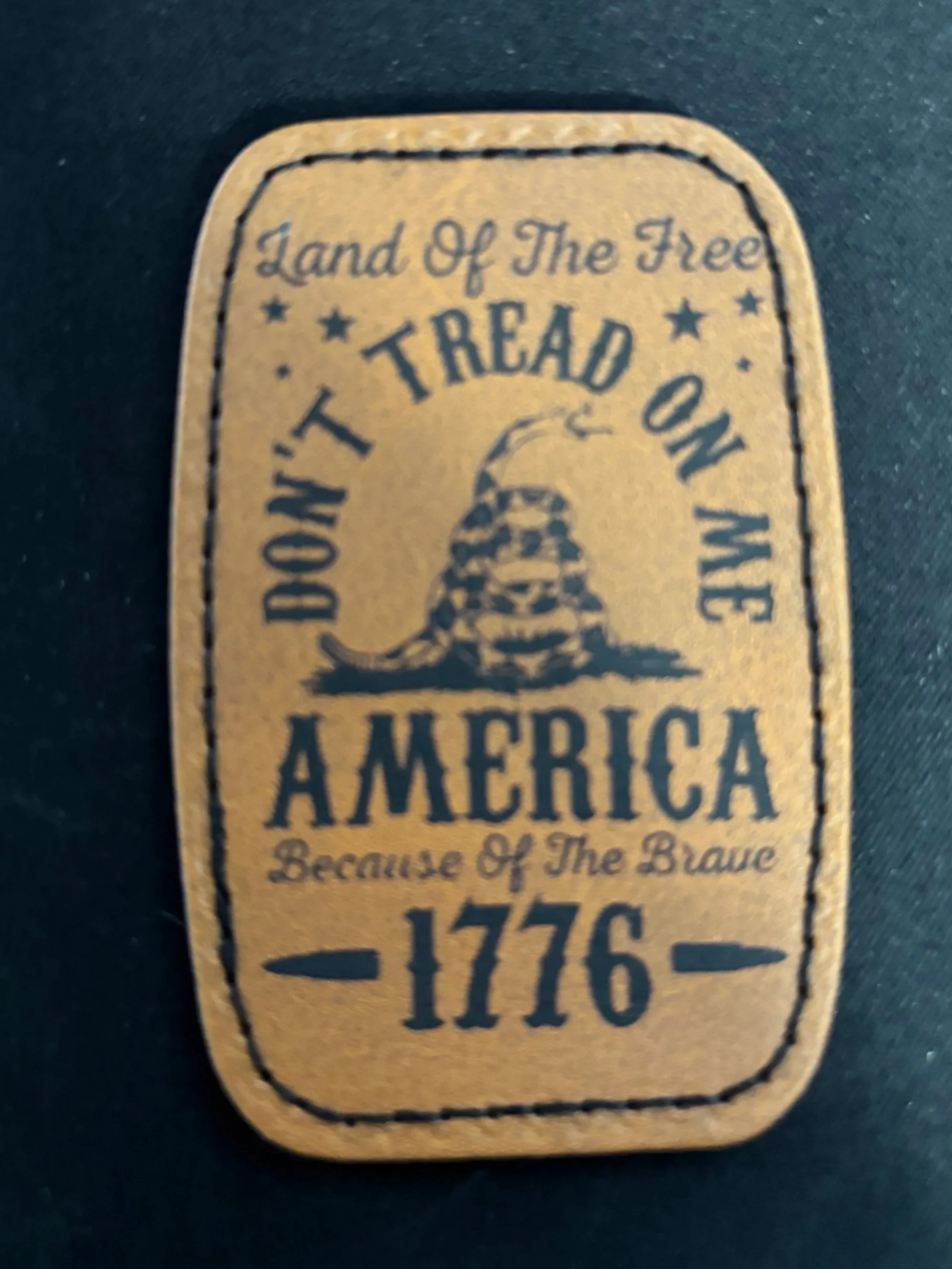 A leather patch with text and a snake graphic, stating "Land of The Free," "Don't Tread on Me," "America," "Because of The Brave," and "1776."