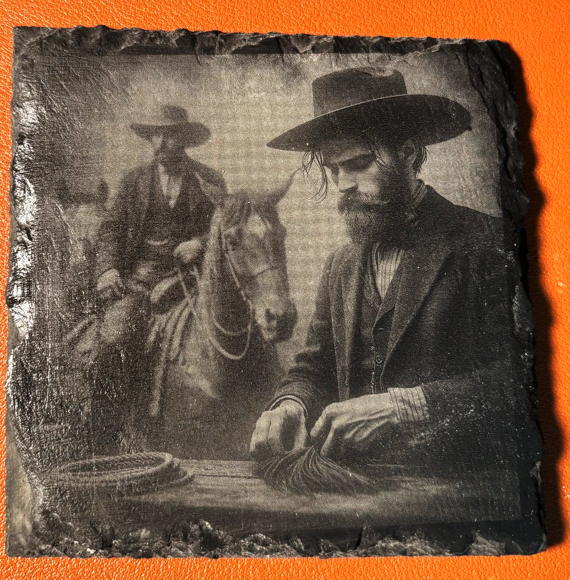A black-and-white photograph of a man with a beard and long hair, wearing a wide-brimmed hat, dressed in vintage clothing, sitting at a table with his hands folded. In the background, there is a person on horseback also wearing a wide-brimmed hat and