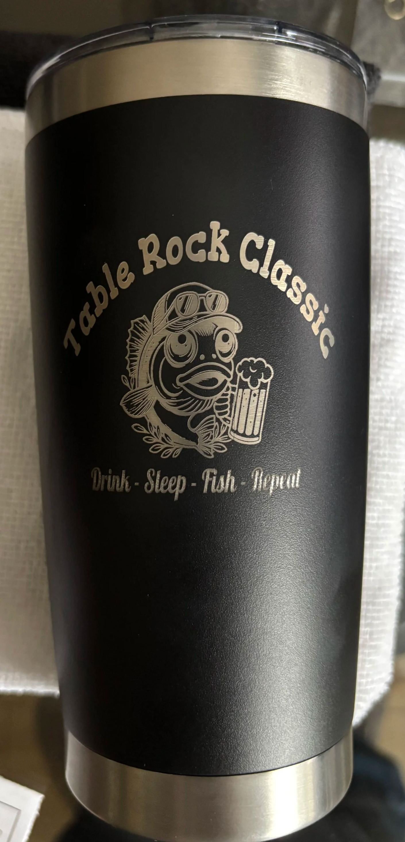 Black travel tumbler with metallic top and bottom, featuring a logo of a fish wearing sunglasses, holding a beer mug, and surrounded by leaves. Text reads 'Table Rock Classic' and 'Drink - Sleep - Fish - Repeat'.