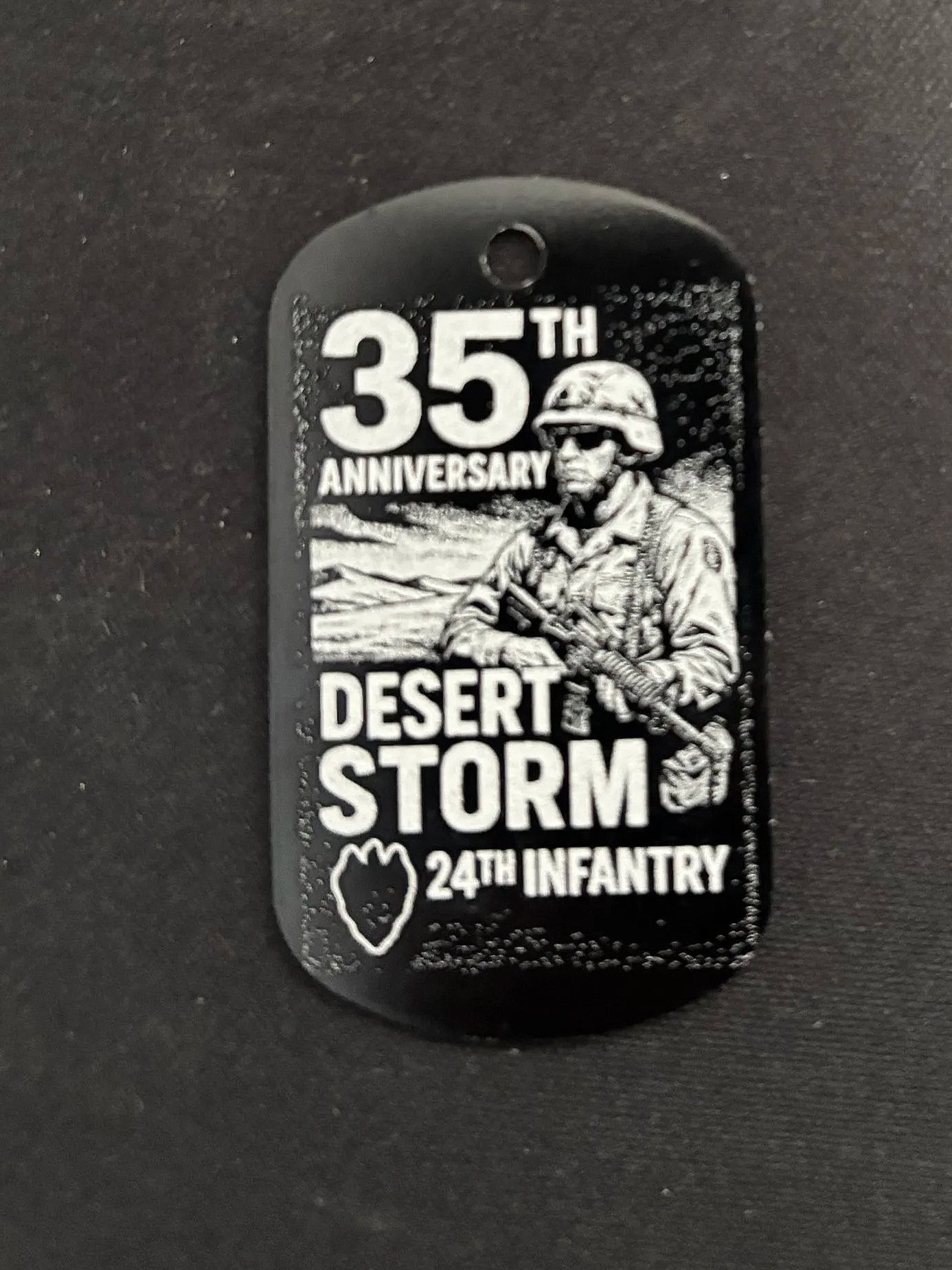 Black and white illustration on a dog tag celebrating the 35th anniversary of Desert Storm, featuring a soldier in uniform wearing sunglasses and a helmet, holding a rifle, with mountains in the background, and a small map of Iraq.