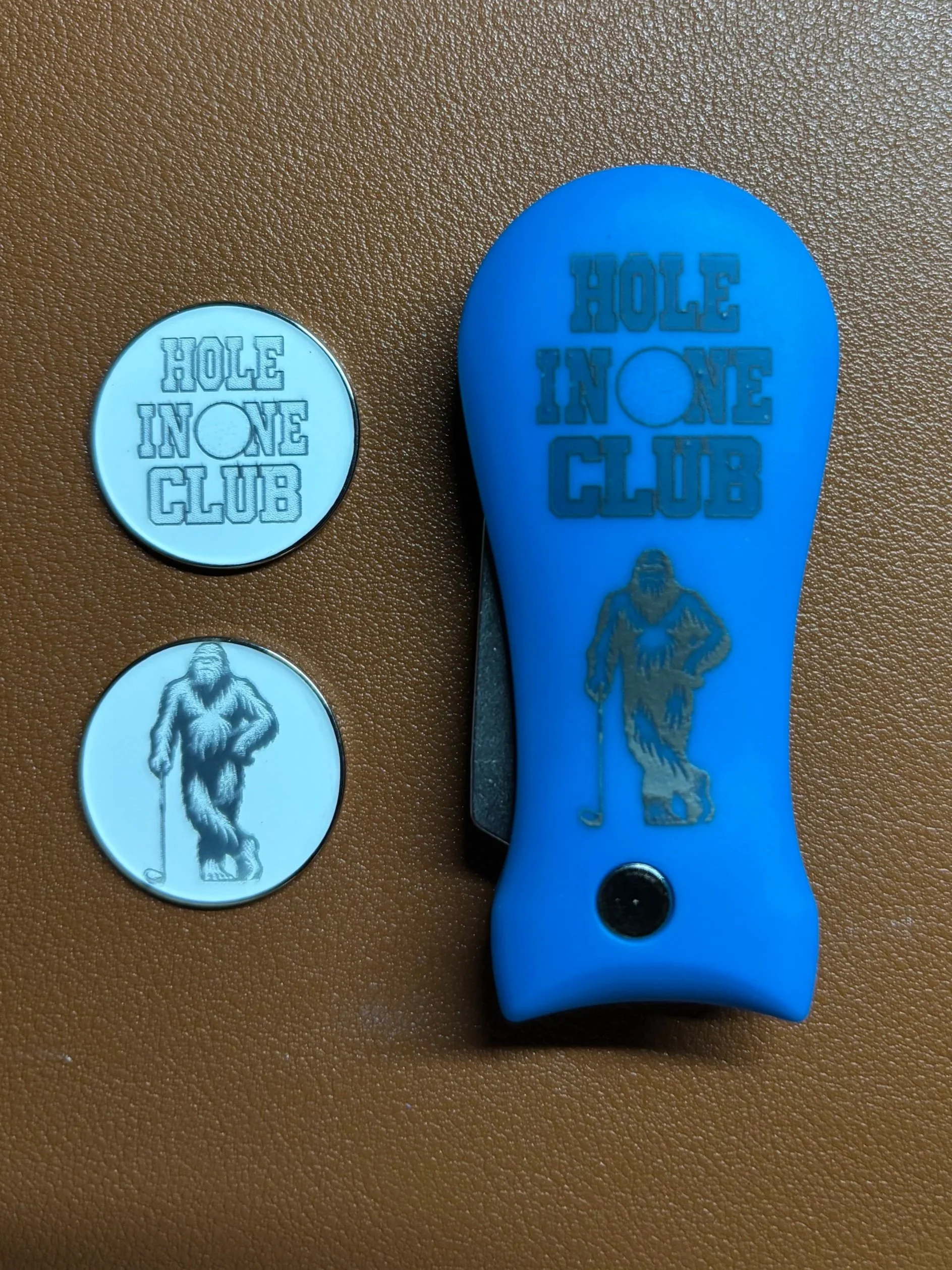 Two round tokens with the logo of the Hole in One Club, one in silver and one in white, and a blue golf divot tool with the Hole in One Club logo and a golfer figure on a brown surface.