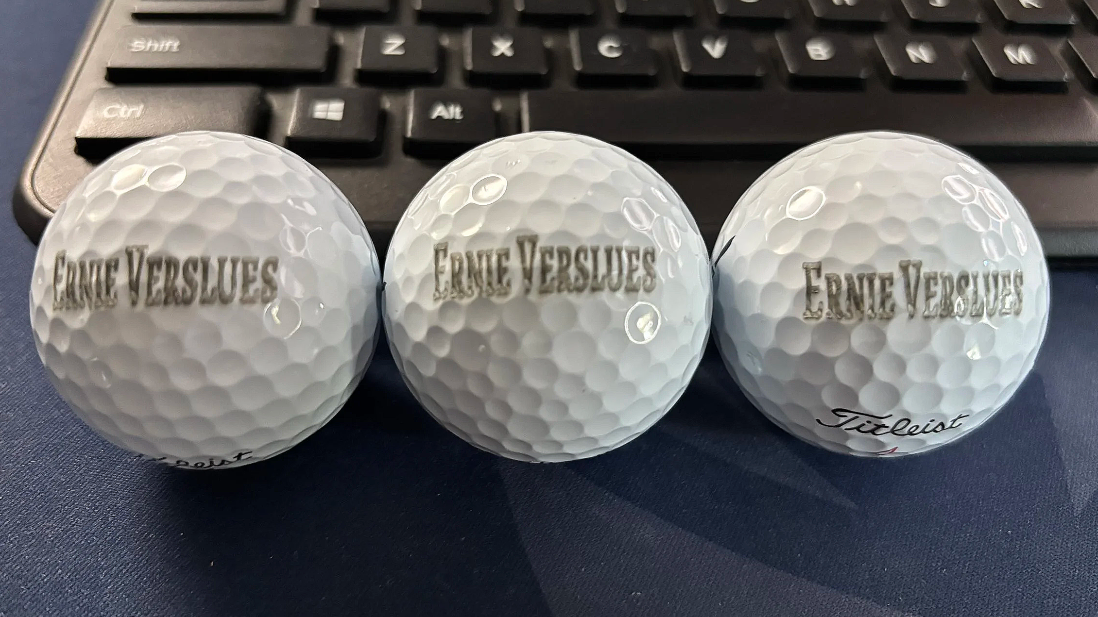 Three white golf balls with 'Ernie Verves' and 'Titleist' written on them, placed on a dark surface in front of a black computer keyboard.