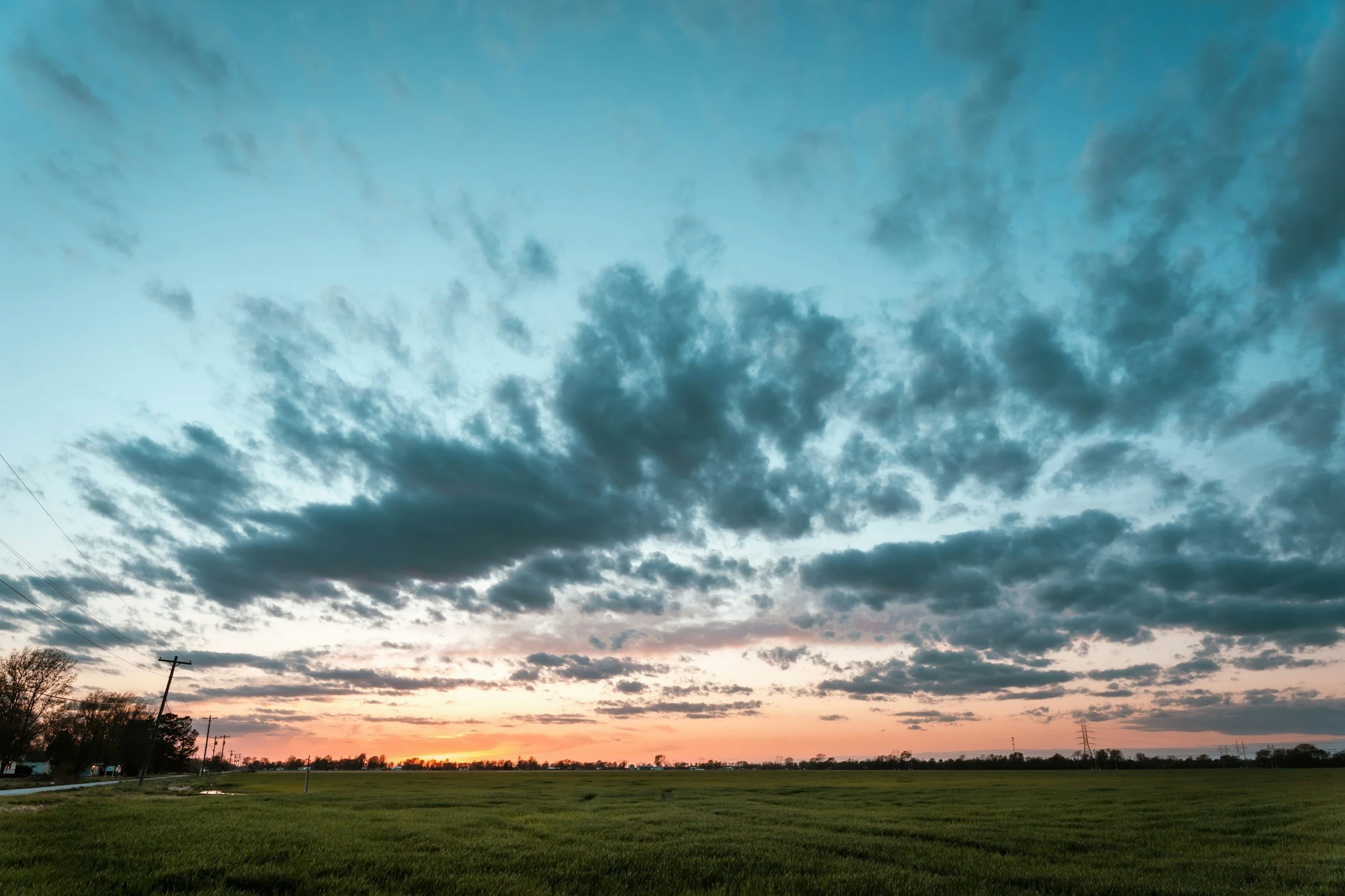 A rural landscape with a green field, a few trees, power lines, and a sunset with a partly cloudy sky.