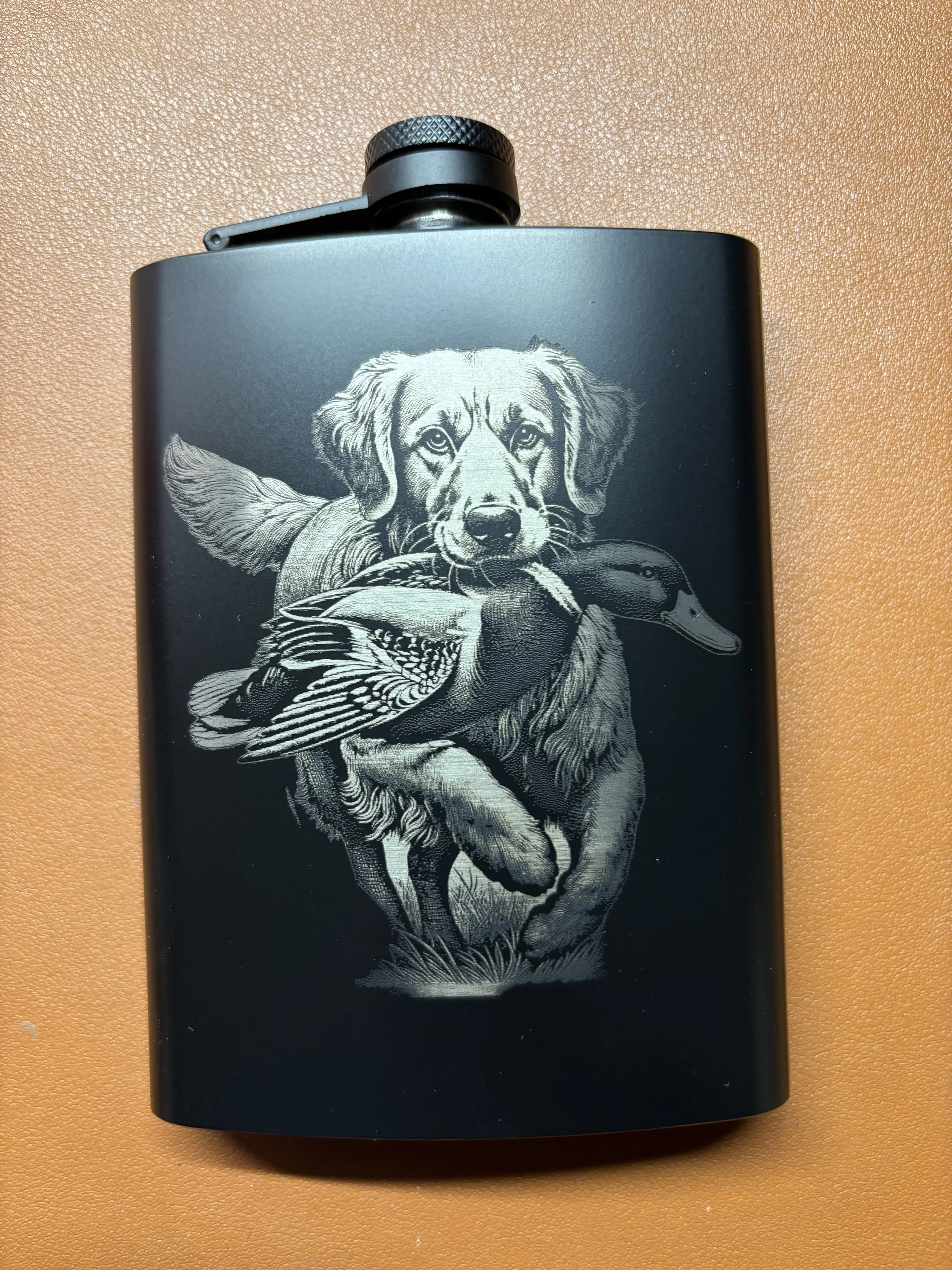 A black flask with a detailed silver engraving of a dog holding a duck in its mouth.