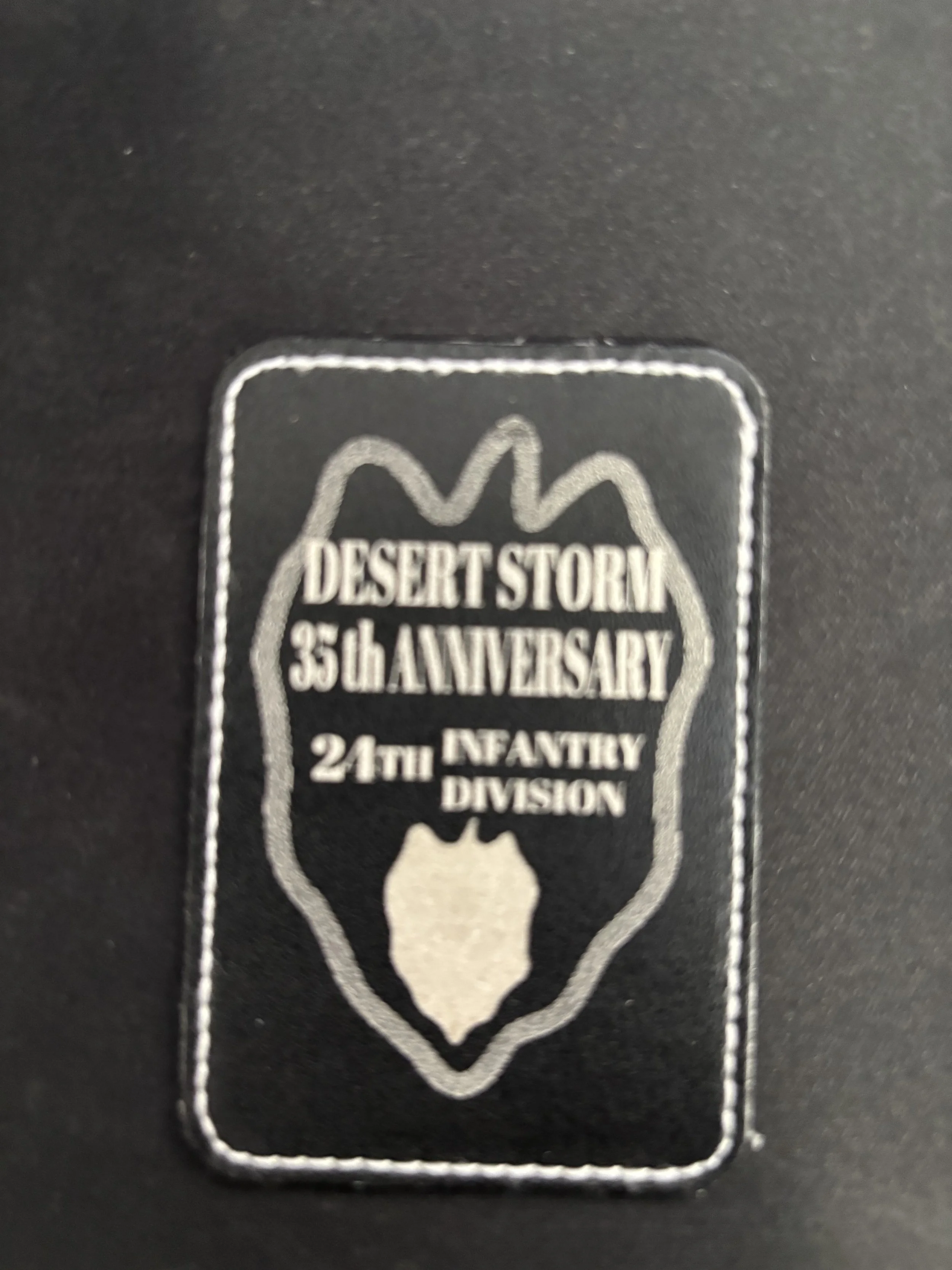 Black fabric patch with silver stitching and text commemorating Desert Storm's 35th anniversary, issued by the 24th Infantry Division, featuring a silver border and a shield emblem in the center.