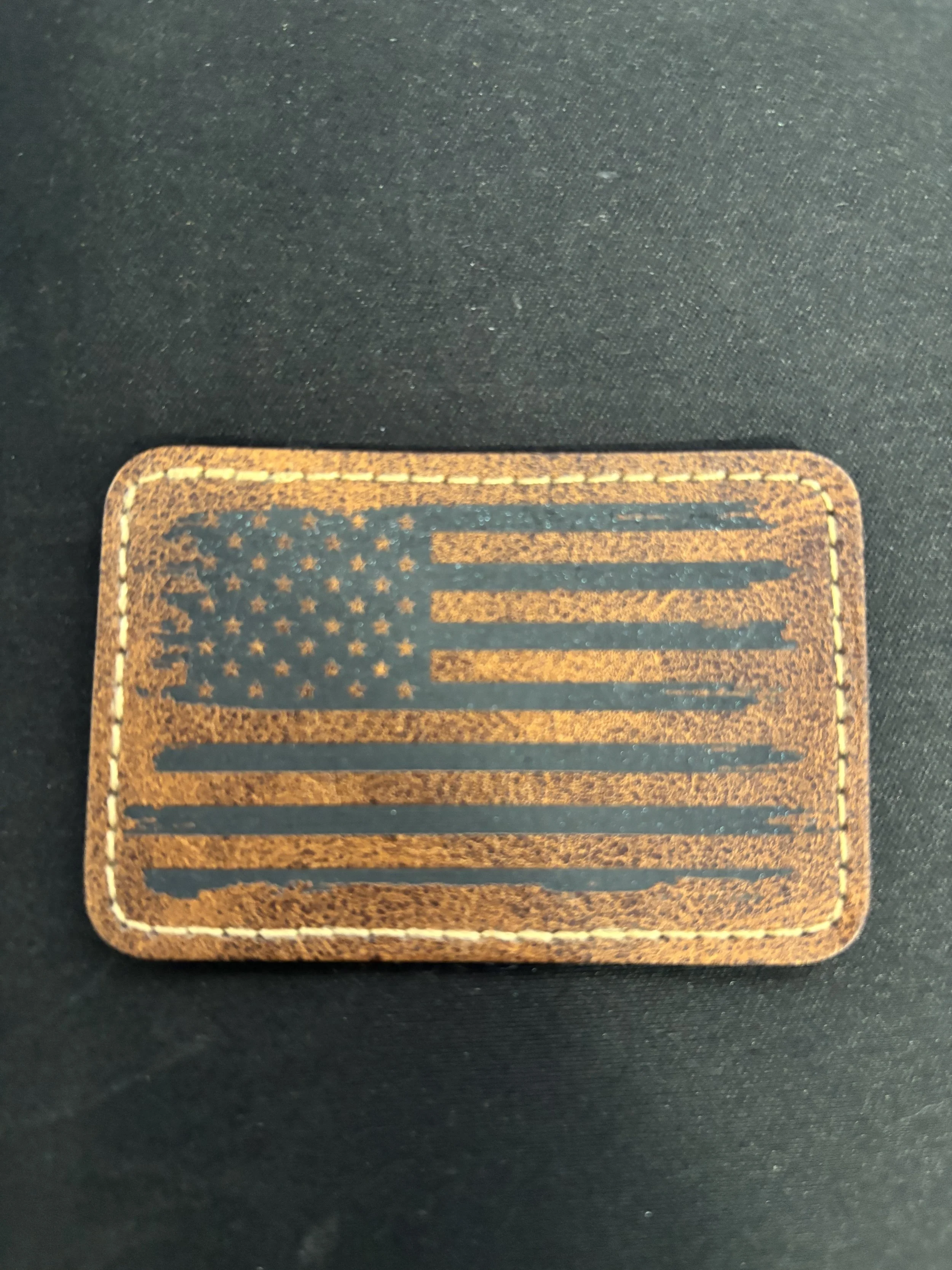 Rectangular leather patch with an American flag design stitched on black fabric.
