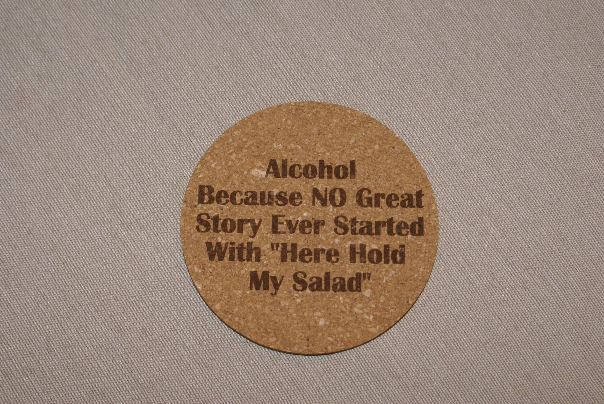 A round cork coaster with humorous text that reads: "Alcohol Because NO Great Story Ever Started With 'Here Hold My Salad'".