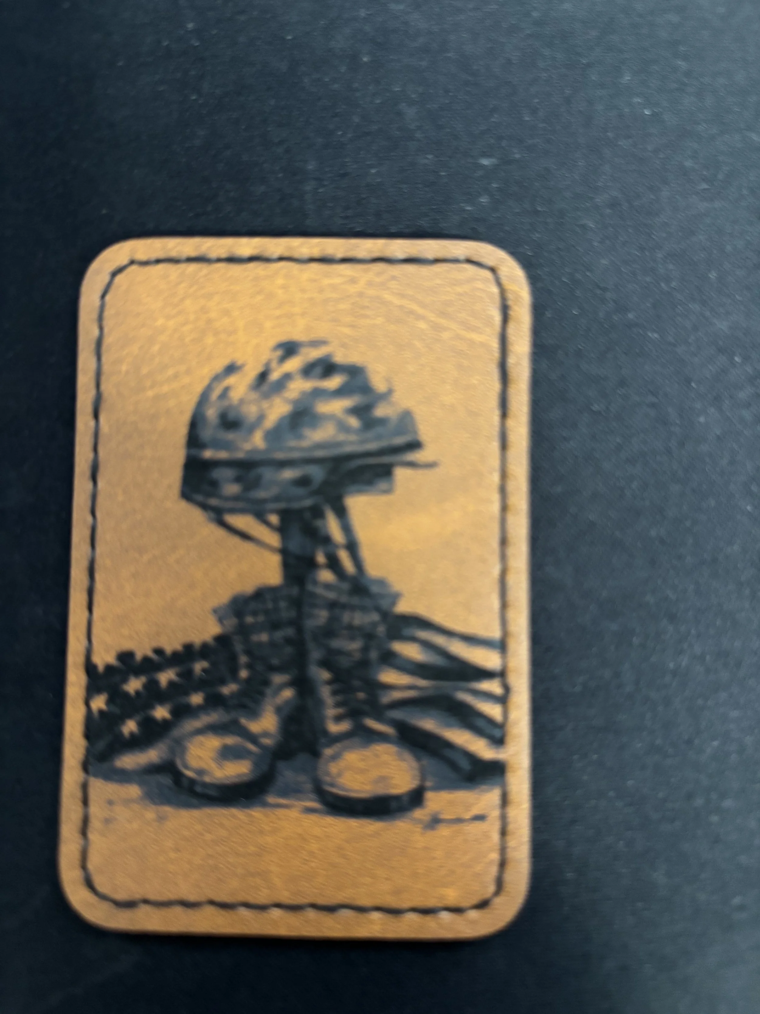 A small rectangular metallic patch with a black drawing of a helmeted soldier standing with two American flags on either side, on a black background.