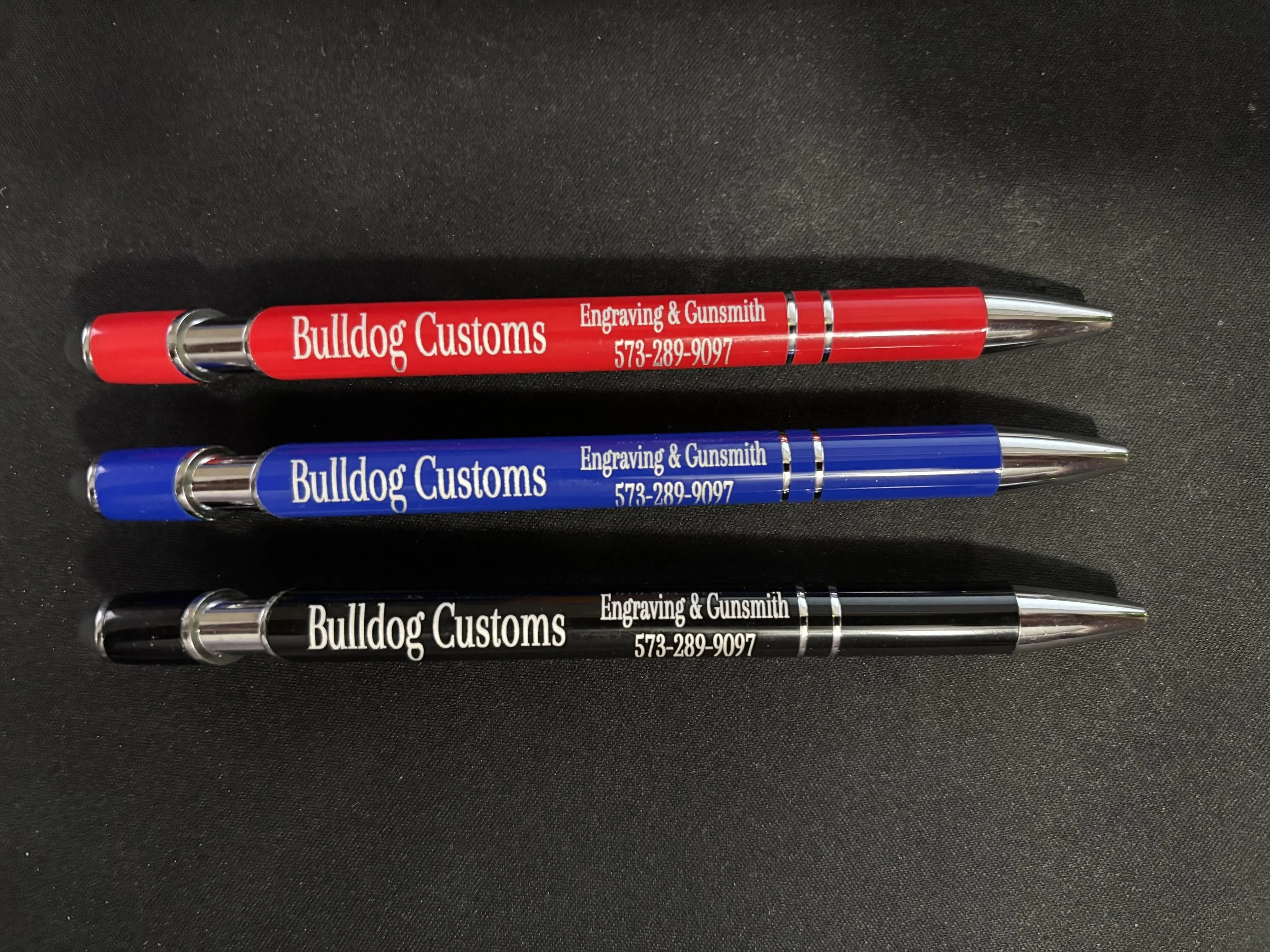 Three custom pens: red, blue, and black, each with white or black text advertising Bulldog Customs engraving and gunsmith services, along with a phone number.