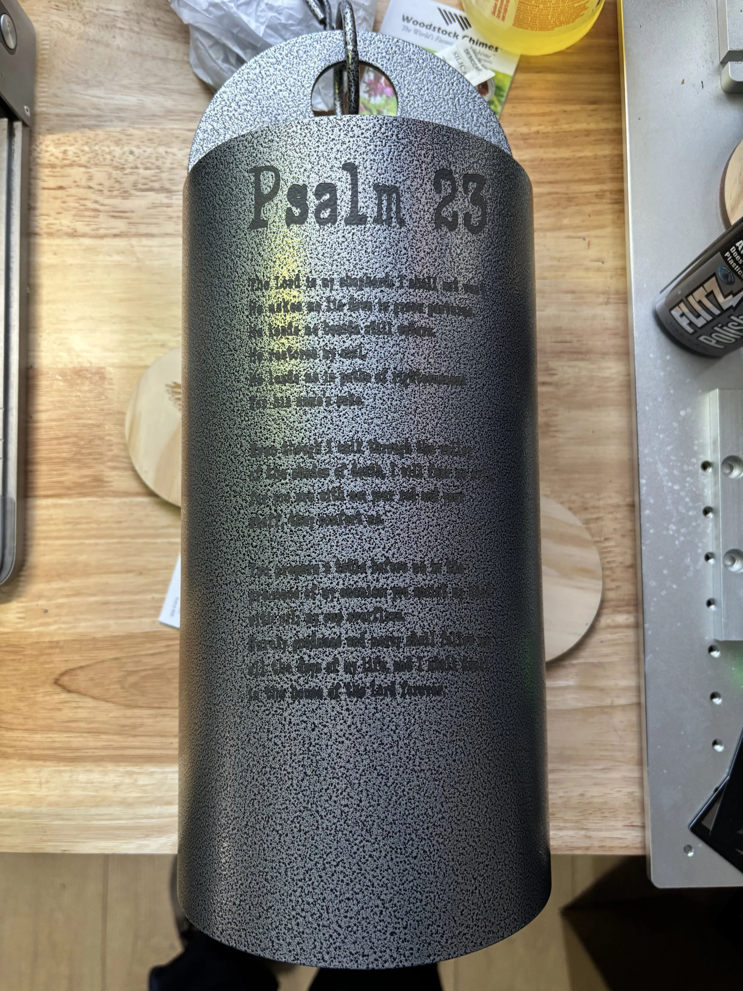 A black cylindrical metal object with a textured grey section at the top that displays the text 'Psalm 23' and a biblical passage engraved on it. The object is hanging on a hook above a wooden surface, with various items around it, including a small 