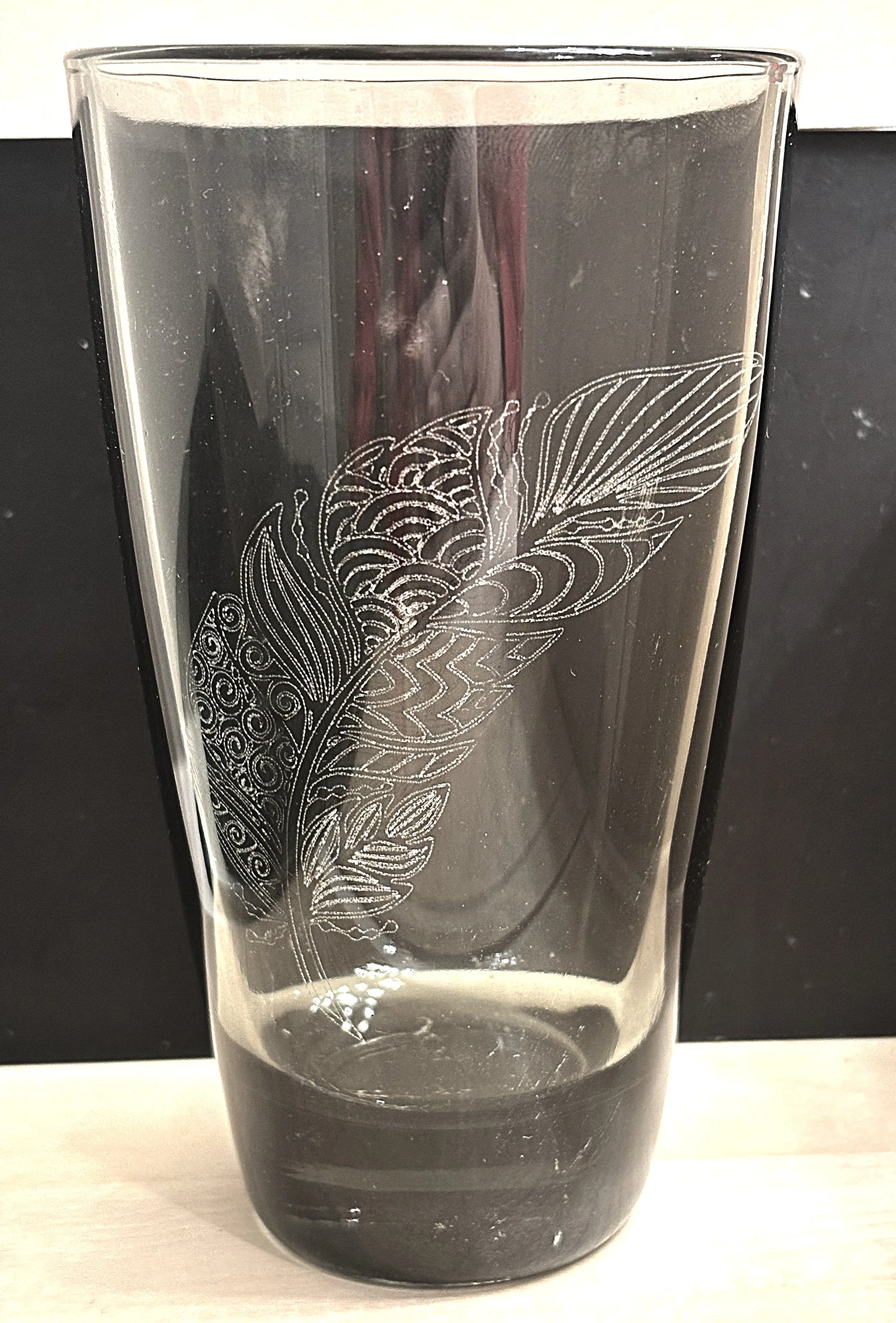 A tall glass with a decorative feather design etched on its surface, placed on a light-colored surface against a black and white background.