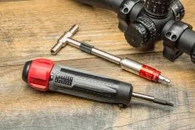 Tools on a wooden surface, including a red and gray screwdriver, a T-handle wrench, and a black scope or binoculars.