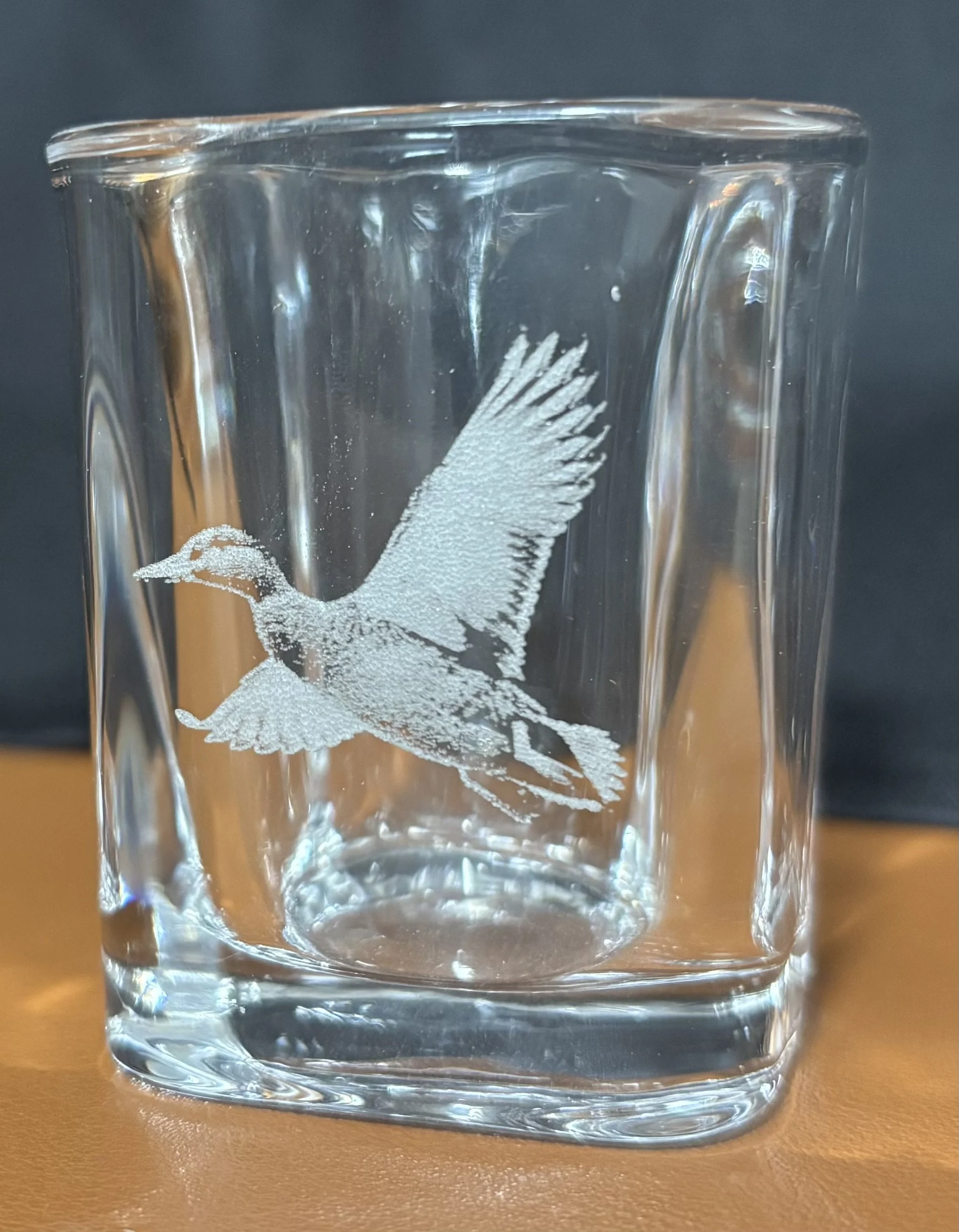 A clear glass tumbler with an etched image of a flying duck on its surface, placed on an orange surface with a dark background.