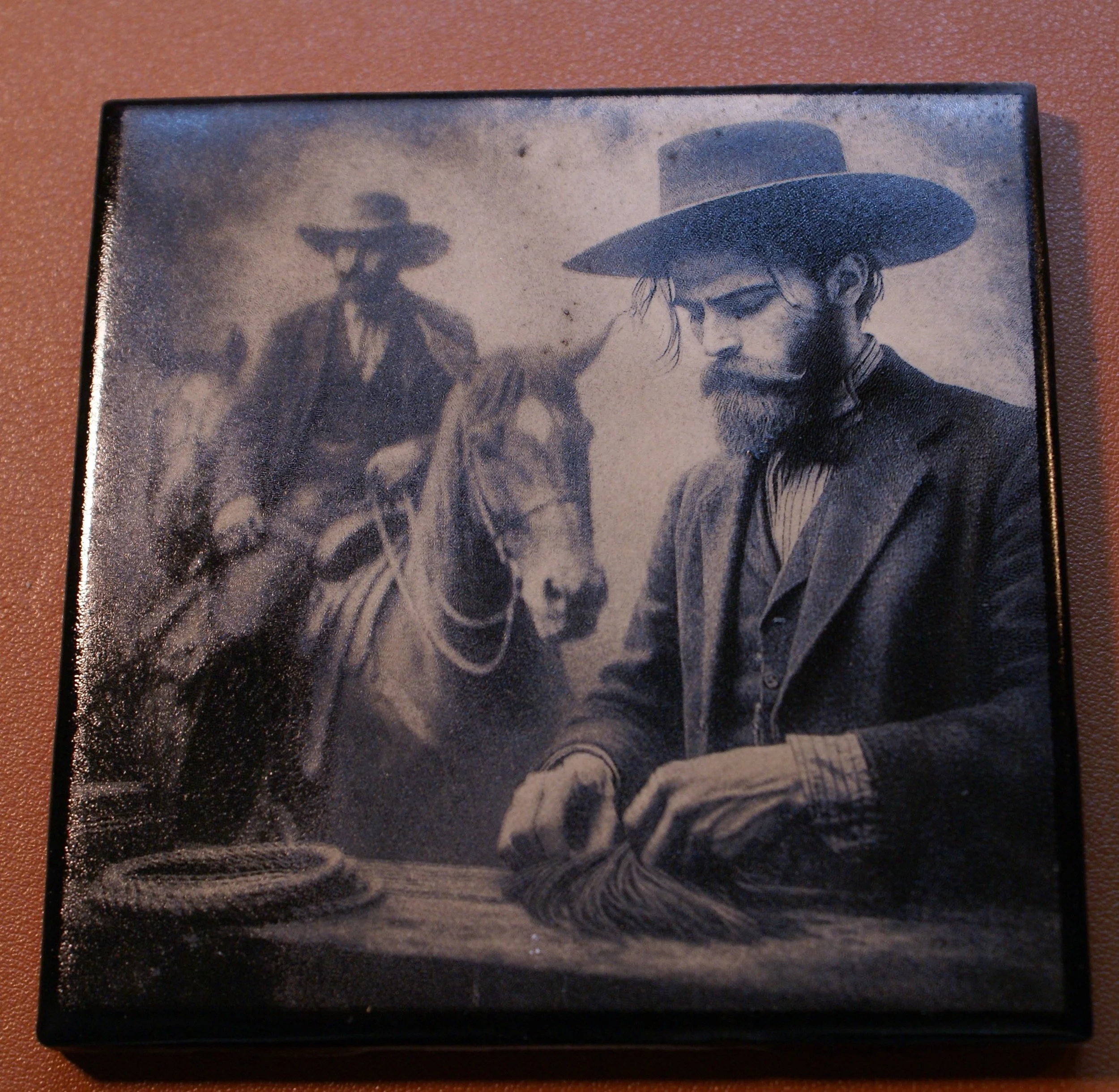 A vintage black and white photograph of a man with a beard wearing a wide-brimmed hat and a suit, sitting at a table with a small rolled item in his hands. In the background, there is a person on horseback wearing a hat, with open landscape surroundi