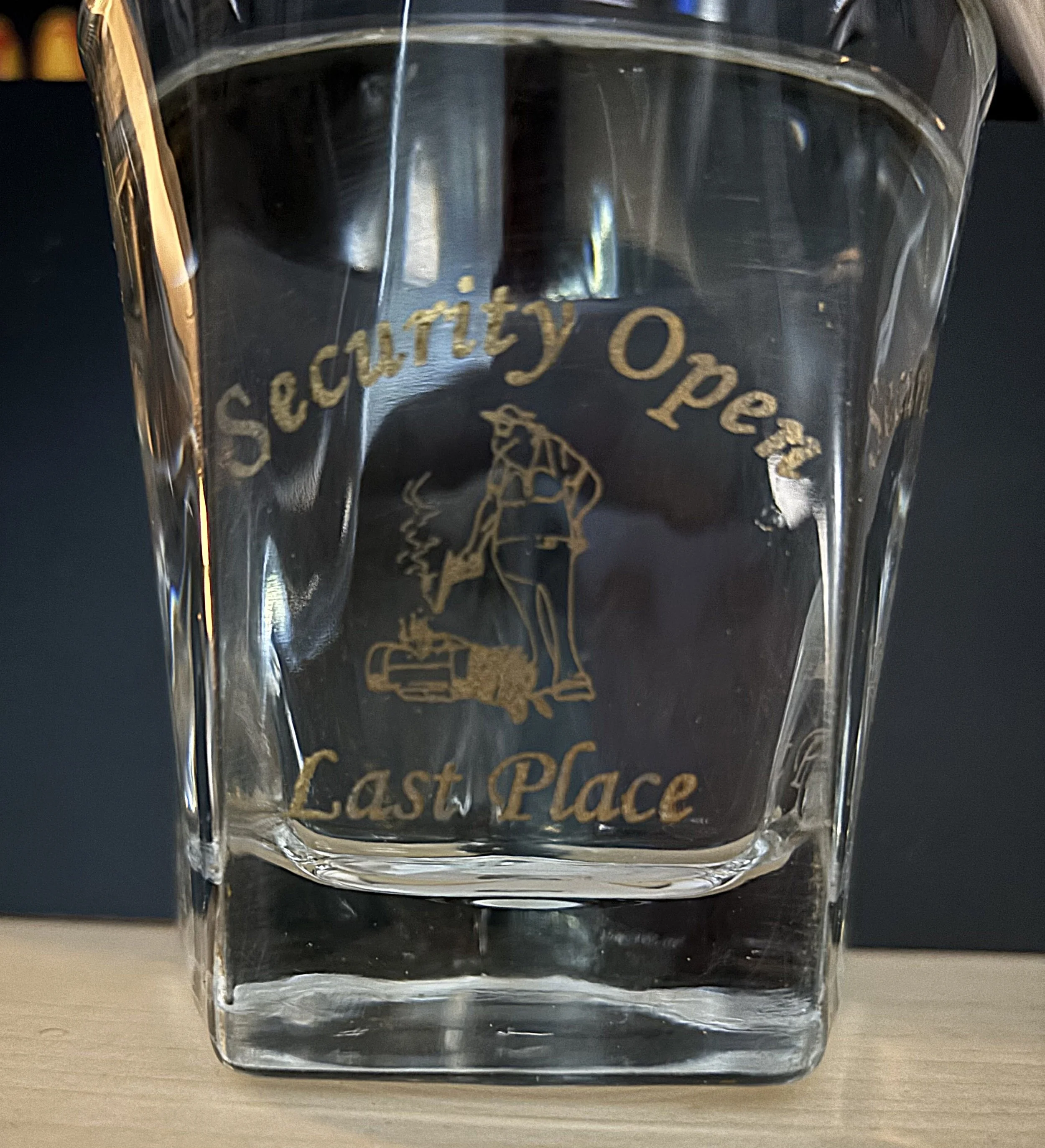 A clear glass with gold-colored text and engraving. The text reads "Security Open Last Place" and features an engraved image of a person leaning on a crate, smoking, with a coffee mug nearby.