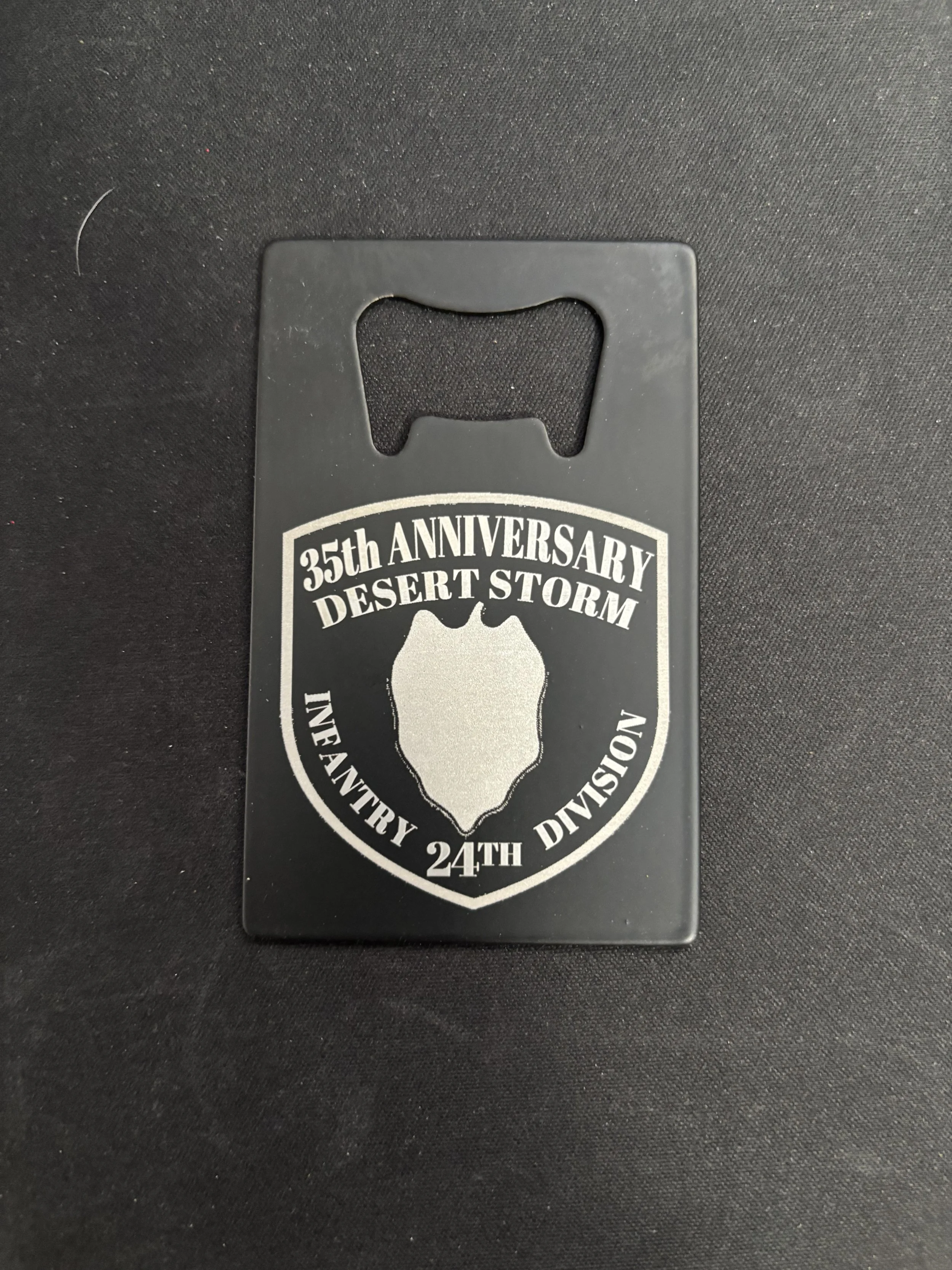 A black plastic bottle opener with a silver emblem commemorating the 35th anniversary of Desert Storm, 24th Infantry Division.